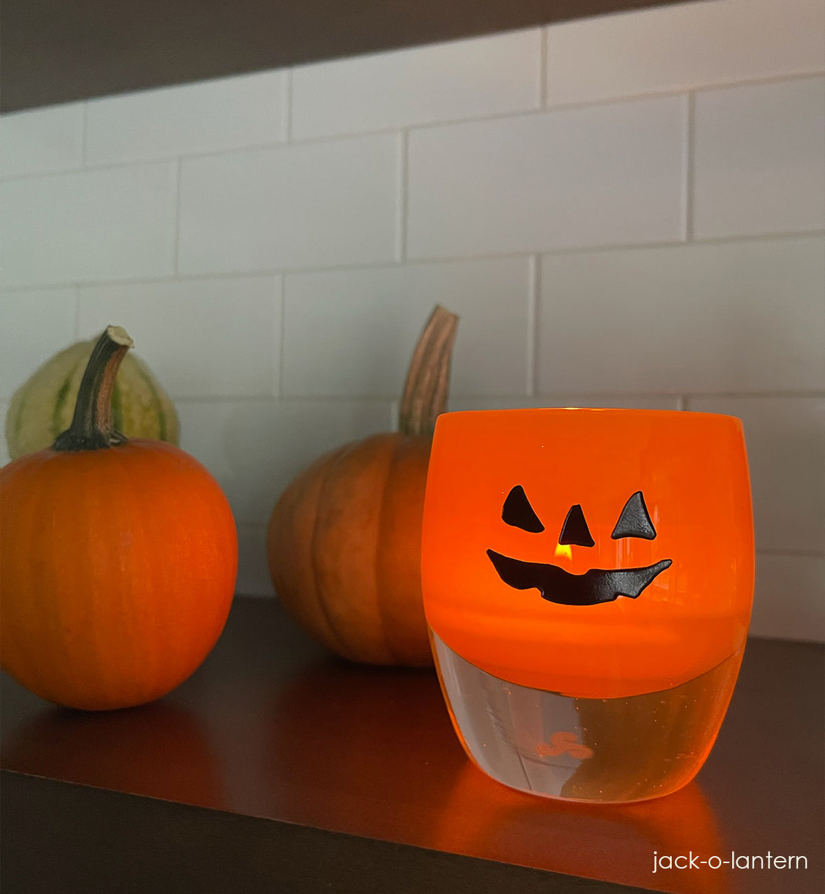 jack-o-lantern, orange hand-blown glass votive candle holder with a Halloween face etched, sitting on a Kitchen shelf with various autumn gords.