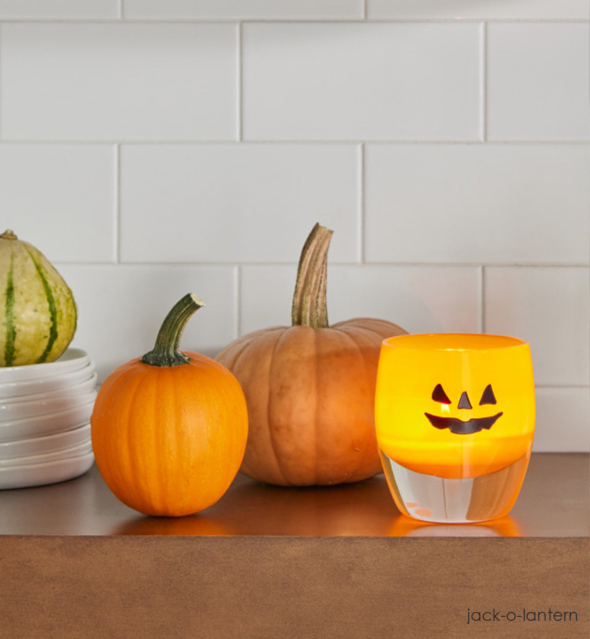 jack-o-lantern, orange hand-blown glass votive with Halloween etched face on side with glowing tealight inside votive. Sitting on brown kitchen shelf with various gords