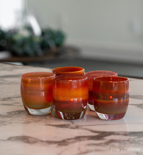 several jane's caramel, caramel brown and purple gradient, hand-blown glass votive candle holders on a black and white marble table with table and plant blurred in background.