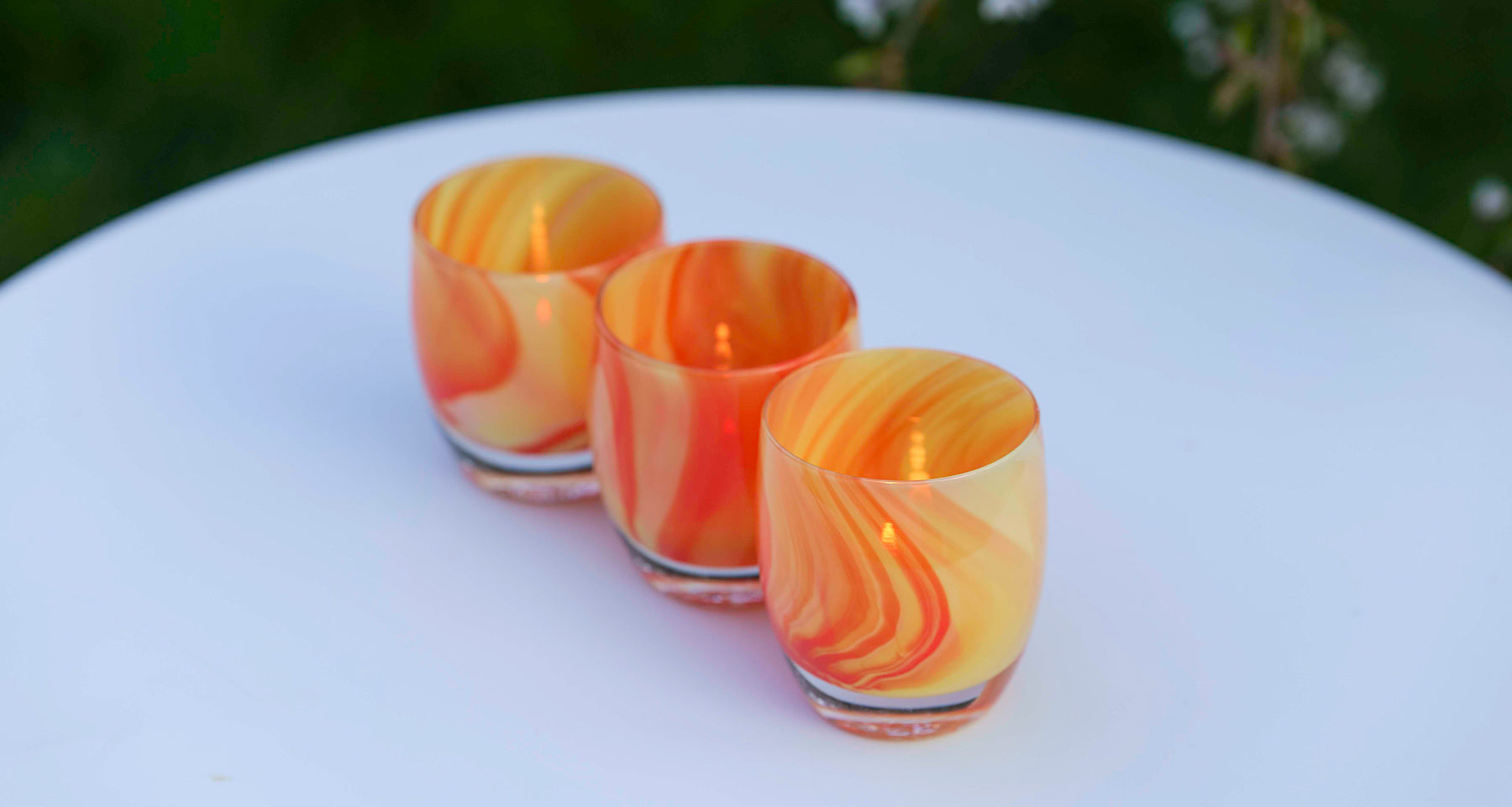 lifesaver, a swirled orange and cream colored hand-blown glass votive candle holder, sitting outside grouped in a line of 3 on a white table.