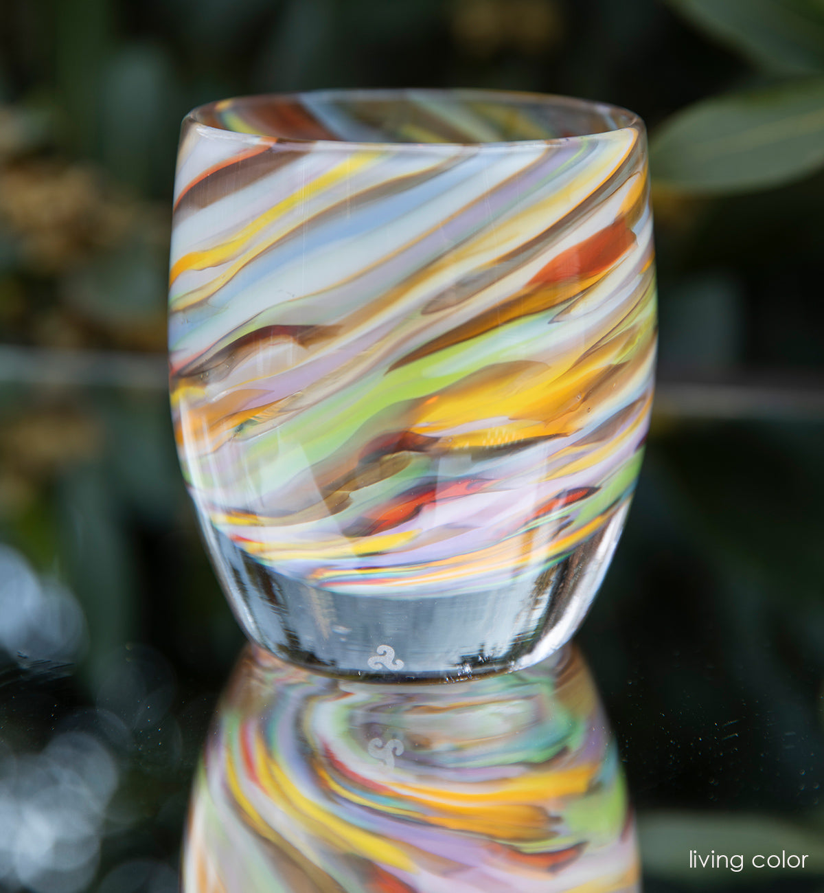 multi-colored blue, brown, yellow, red, purple, green swirl hand-blown glass votive candle holder sitting on a reflective table outside. 