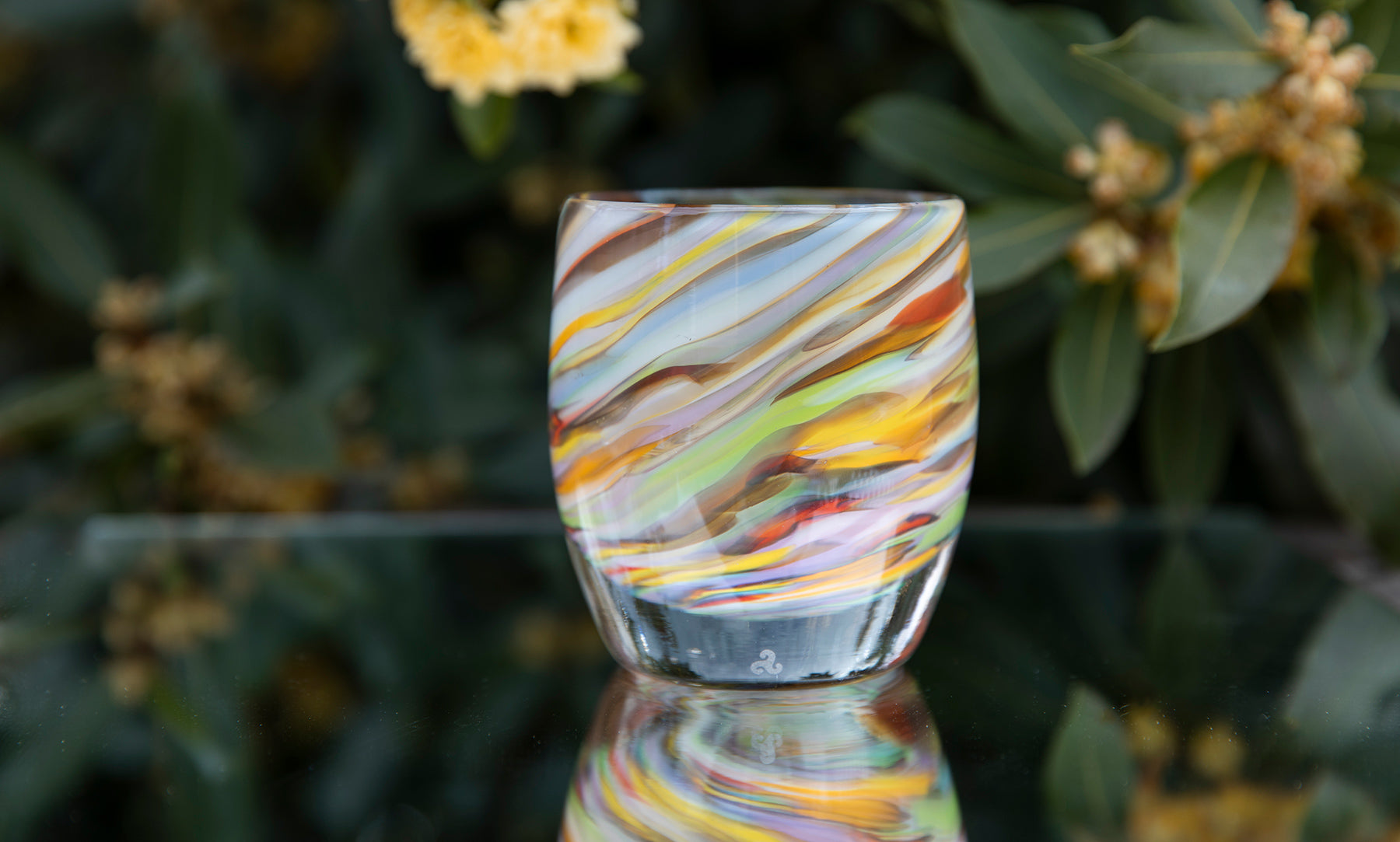 living color, hand-blown glass candle holder with multiple colors resting on a glass table with flowers in the background. 