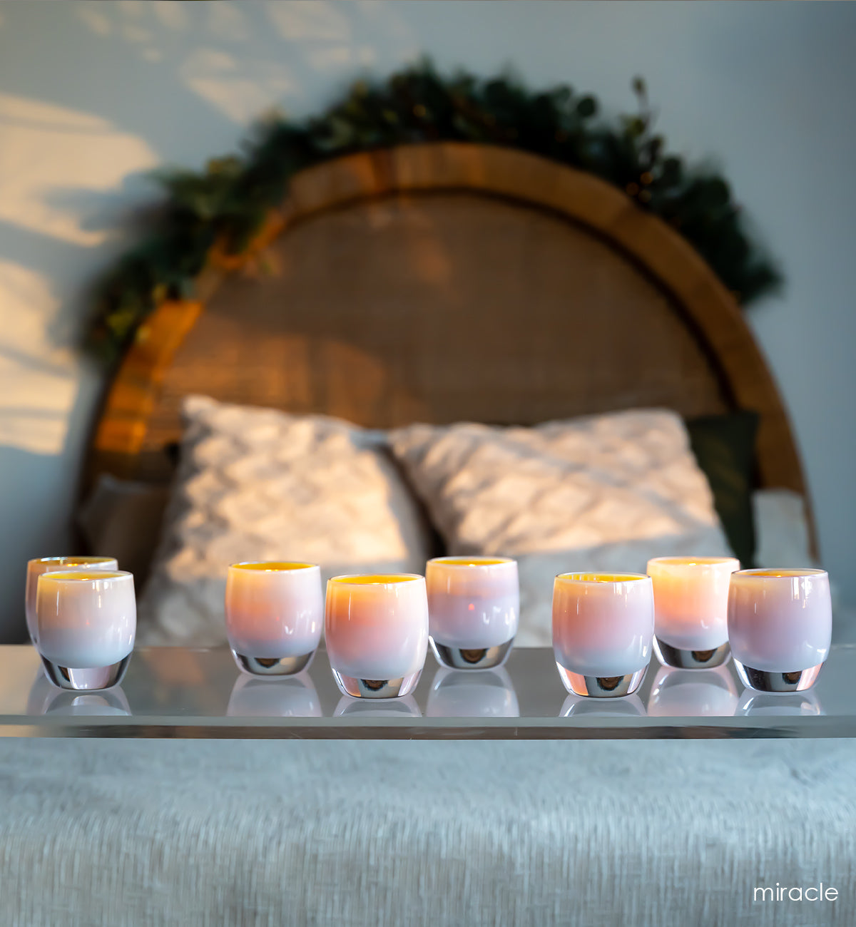 several miracle, golden pink hand-blown glass votive candle holders on a reflective surface with boho bed behind.