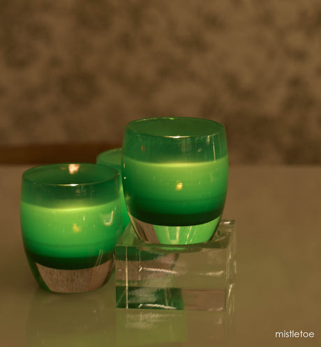 three mistletoe green hand-blown glass votive candle holders, one on a square baby stand next to two on a white table in front of green patterned wallpaper.