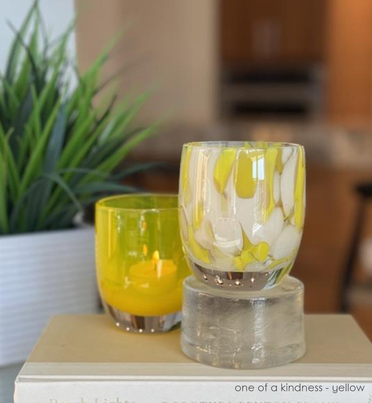 Two hand-blown glass holders, one yellow and one with yellow and white design sitting on books, with a blurred background.