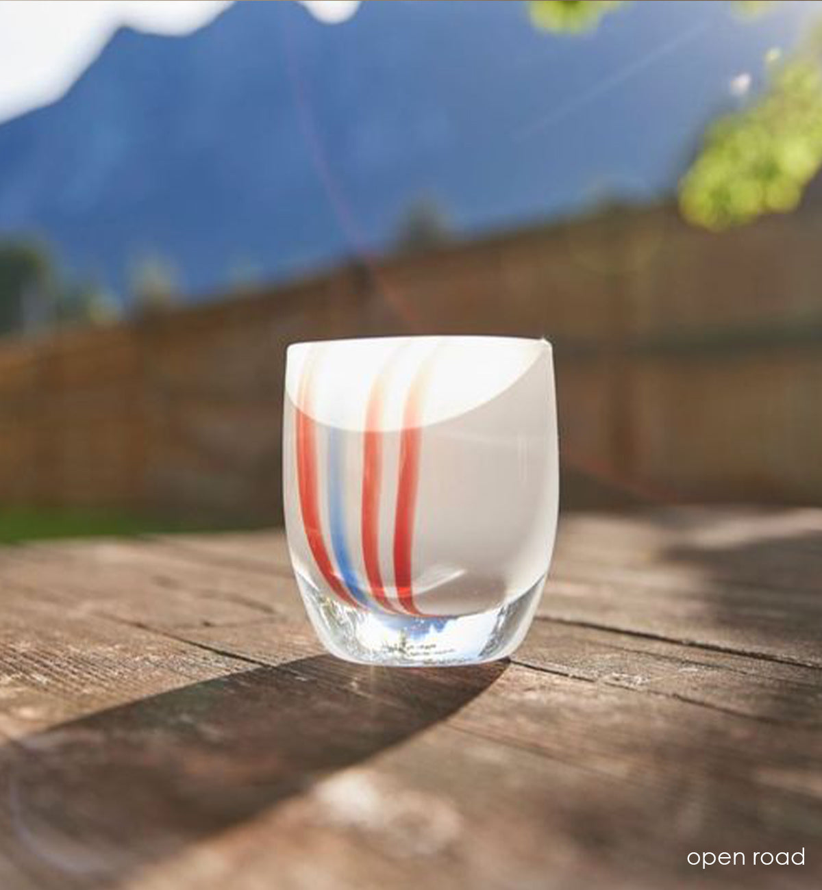 open road, white with red and blue vertical striped hand-blown glass votive candle holder sitting in sunlight on a wood table with leaves and mountains in background.