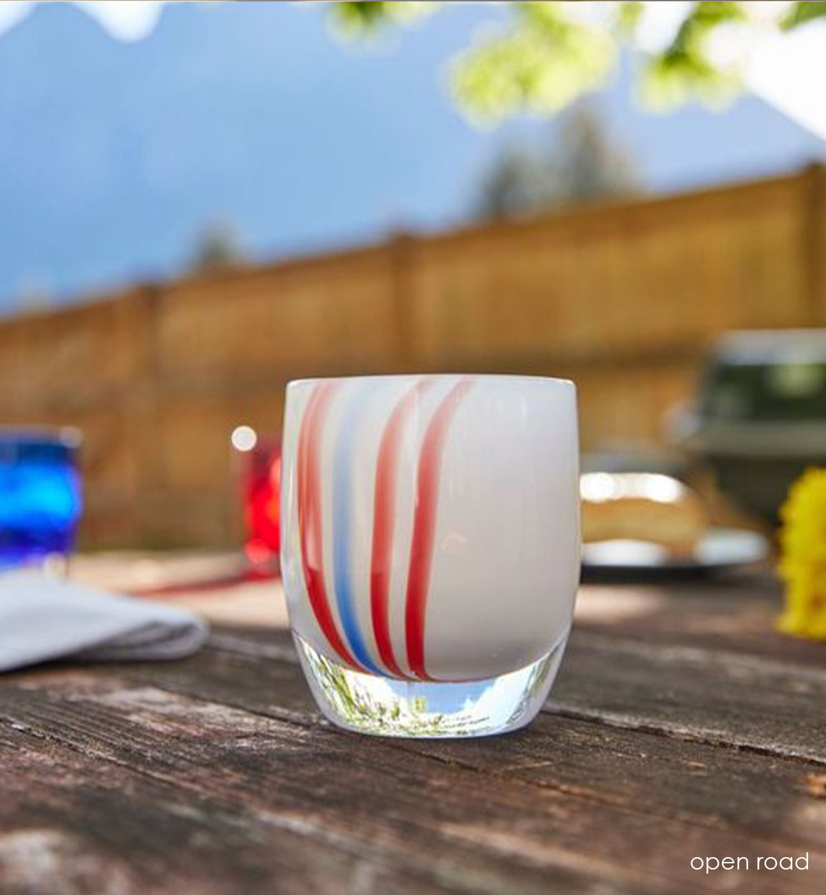open road, white with red and blue vertical striped hand-blown glass votive candle holder on a wood picnic table with red and blue rockers and yellow flowers blurred in background.