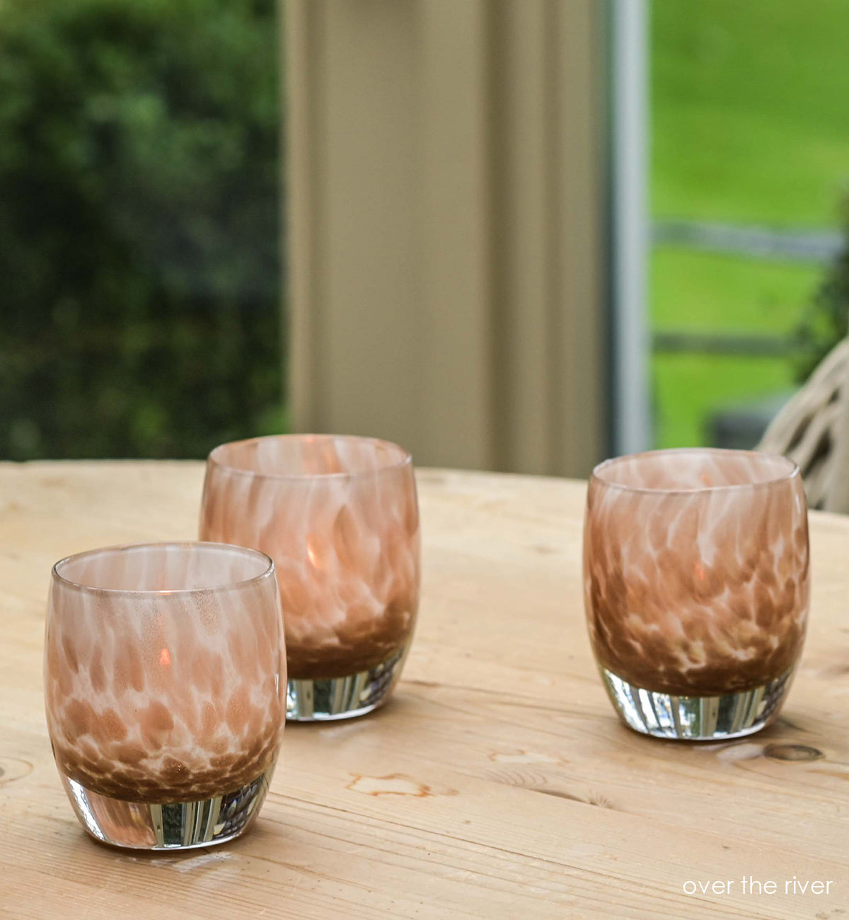 three over the river, gold glitter swirled petals, hand-blown glass votive candle holders on a light wood circular table with greenery outside the windows in background.