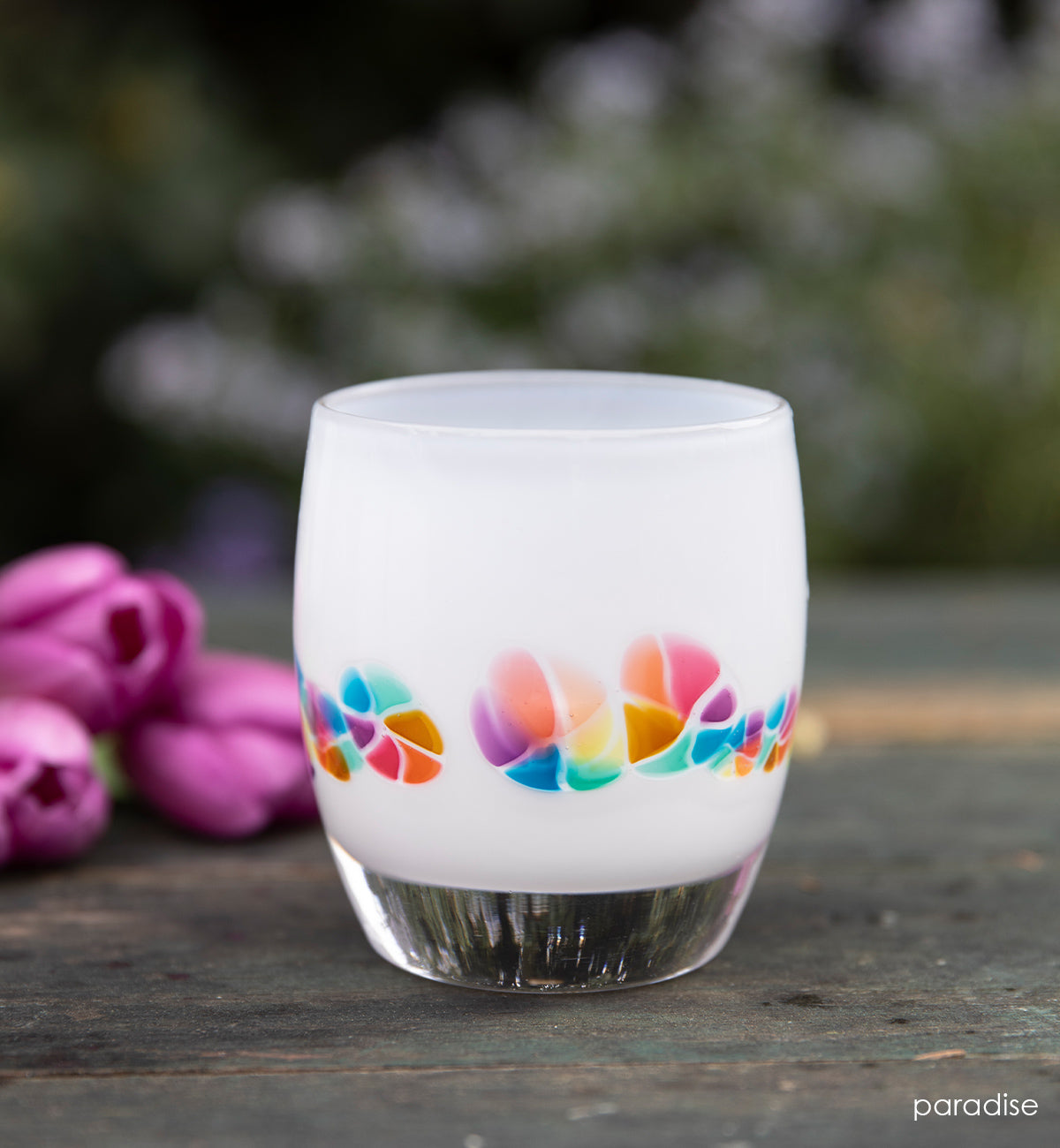 paradise white rainbow murrini detail hand-blown glass votive candle holder on a wood surface outside next to pink tulips.