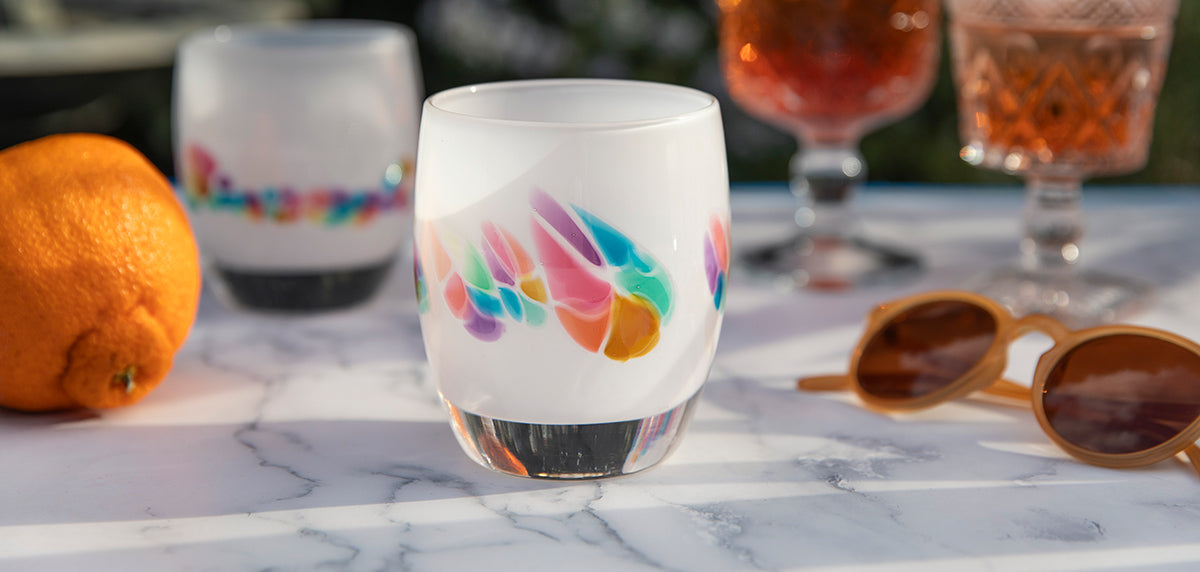 paradise white rainbow murrini detail hand-blown glass votive candle holder on a white surface with sunglasses, an orange and 2 drinks..