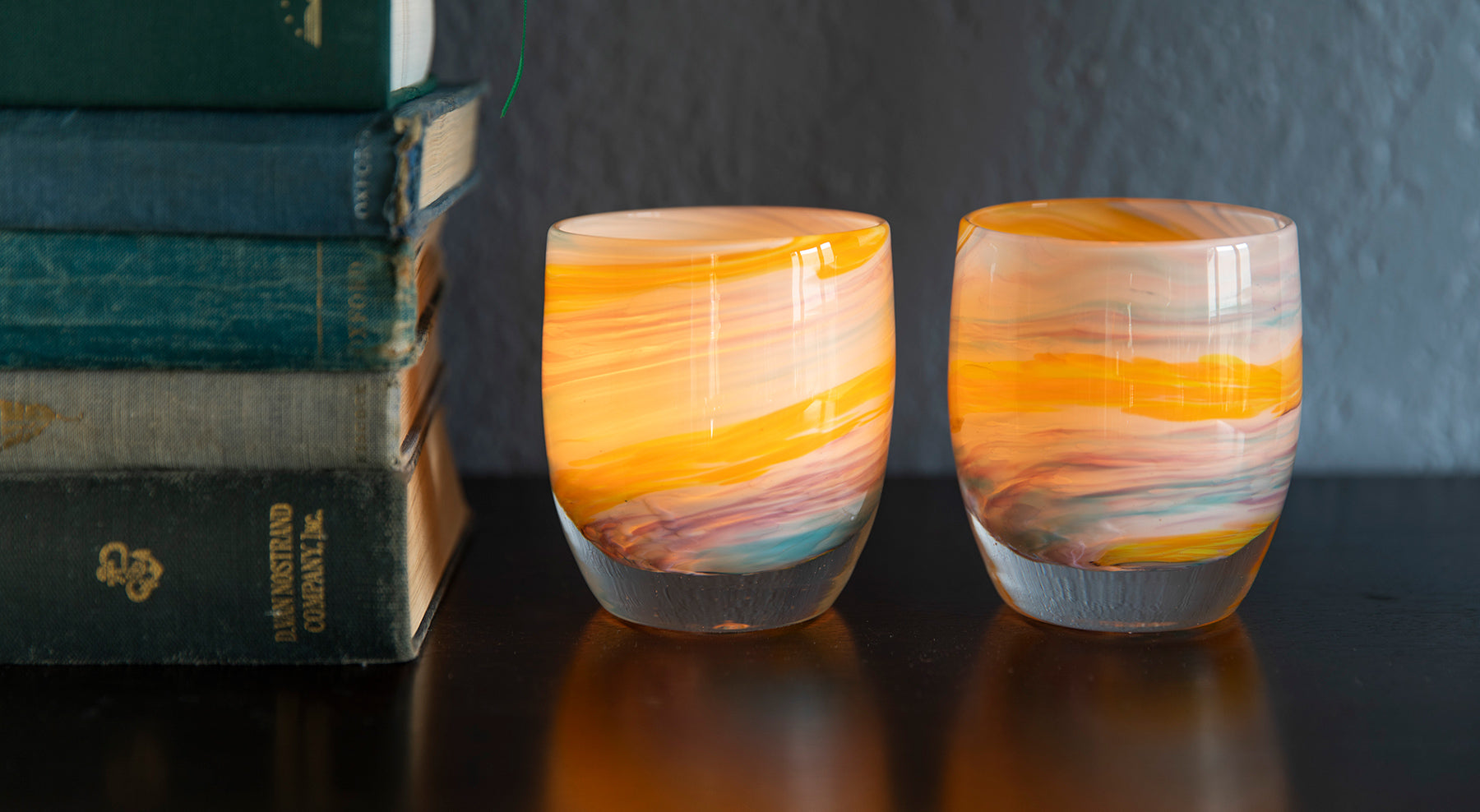 two places you'll go multi-colored yellow, white, purple, blue hand-blown glass votive candle holders sitting on a dark wood table with a stack of books.