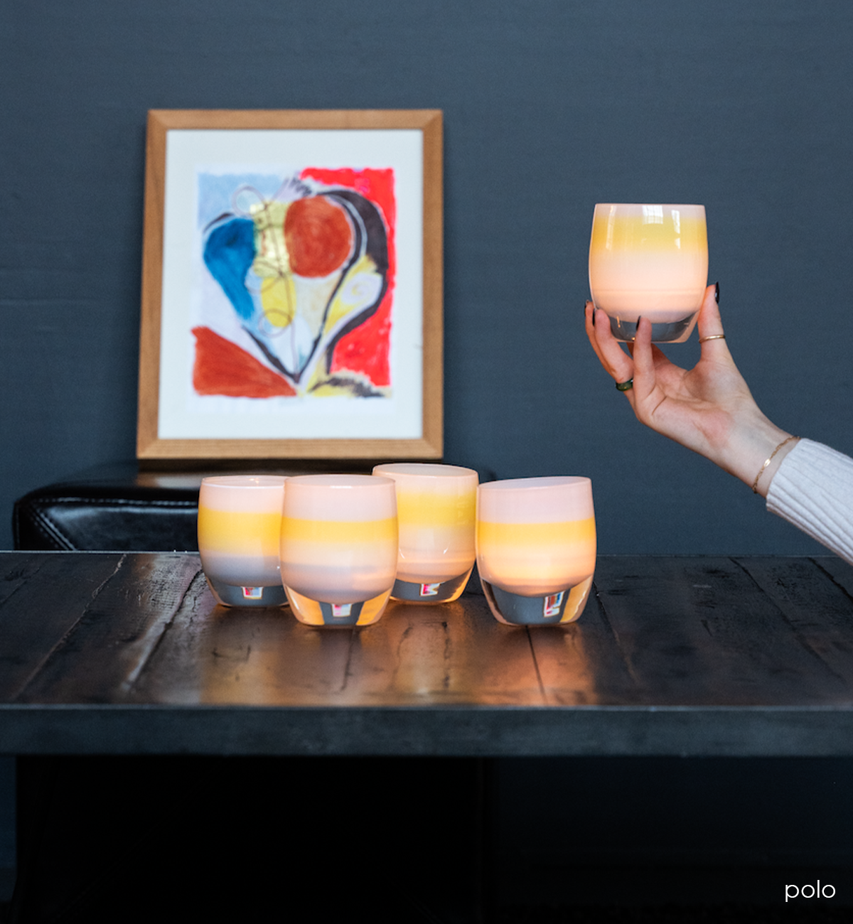 several polo light blue with yellow band in the center hand-blown glass votive candle holders, one of which being held, the rest on a dark table in front of a colorful art piece.