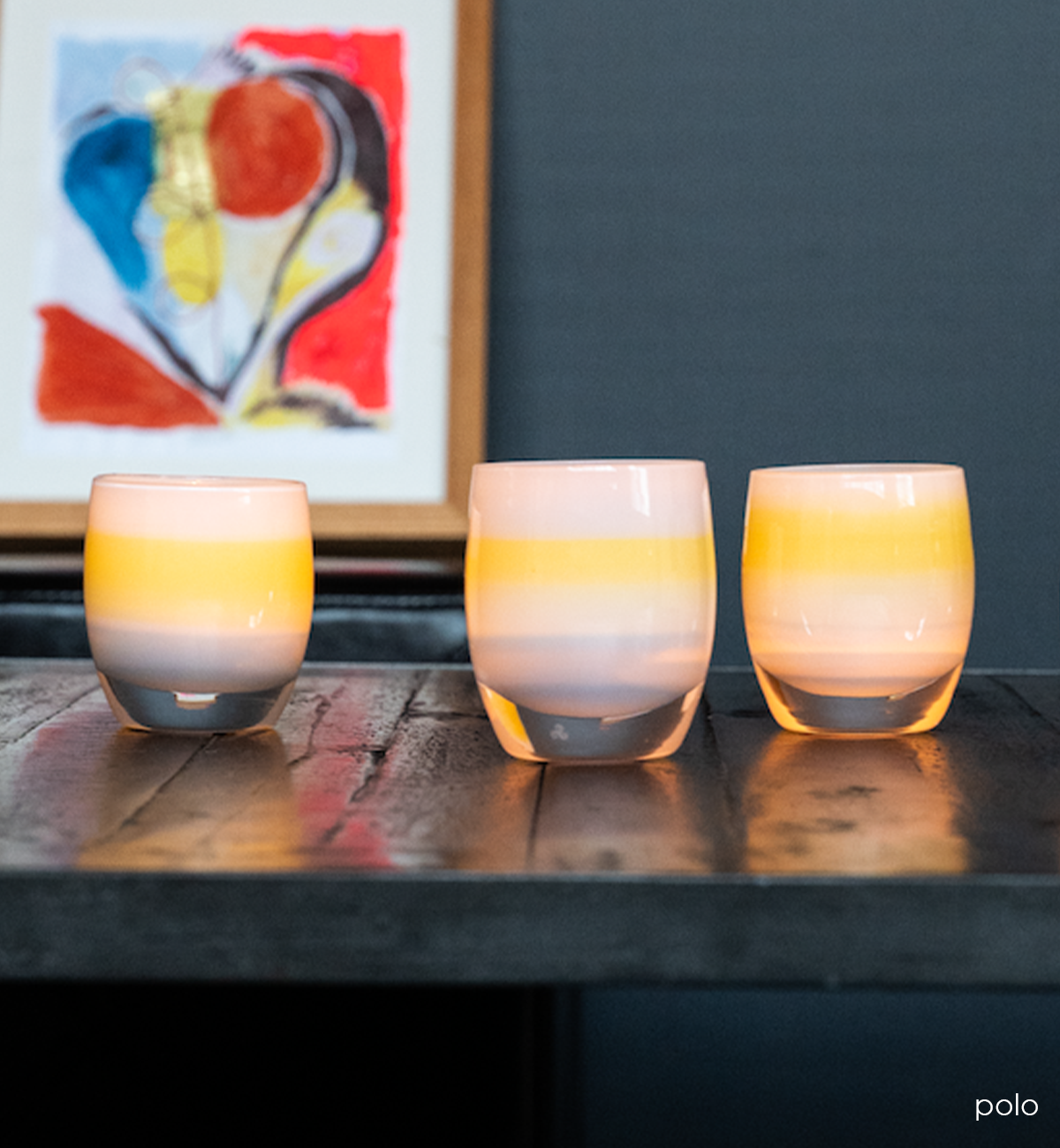 three polo light blue with yellow band in the center hand-blown glass votive candle holders on dark table in front of colorful art piece.