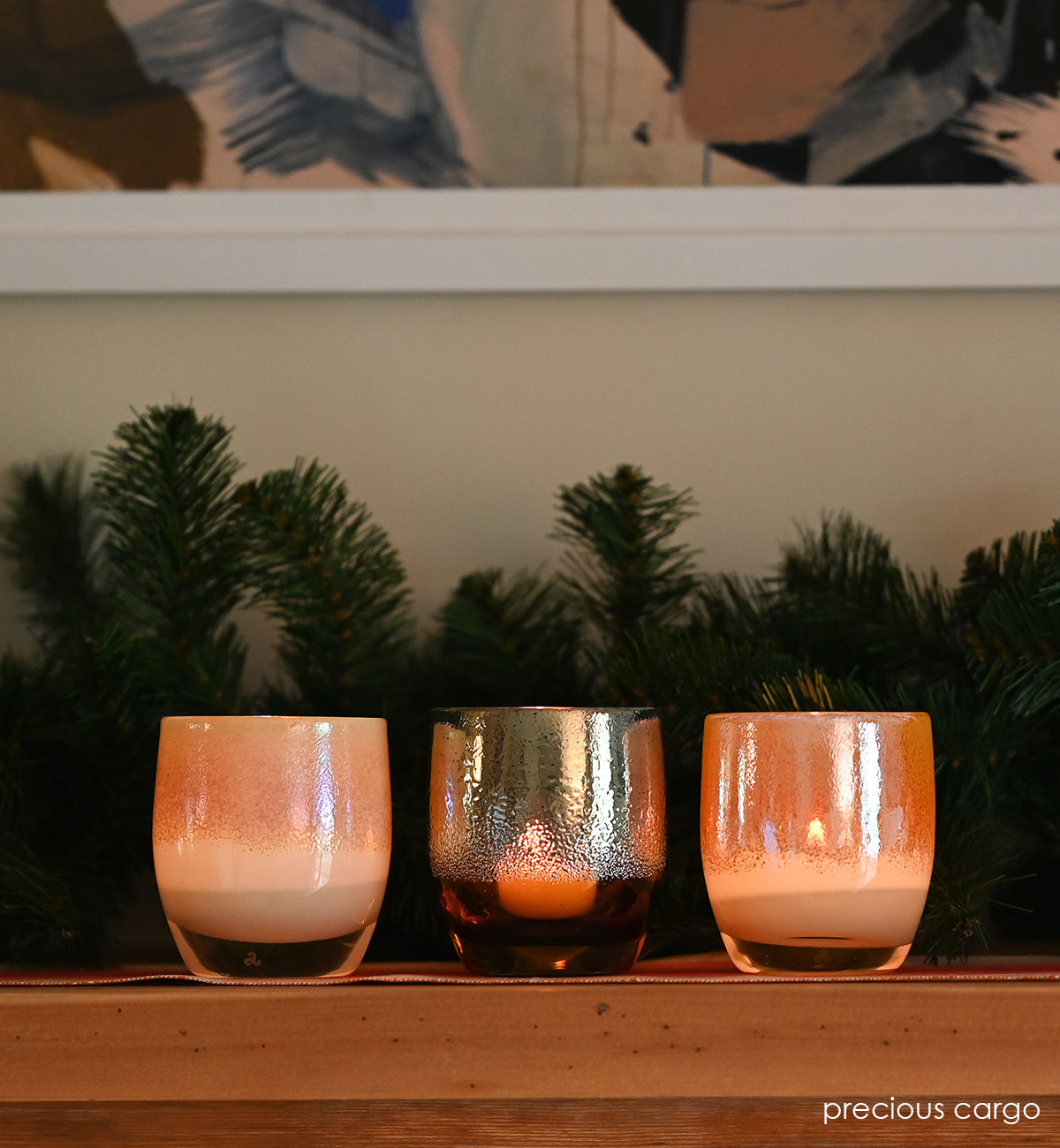 precious cargo is a beautiful set of 3 hand-blown glass votive candle holders; frankincense, gold, and myrrh. sitting atop a light wood mantle with framed wall art and decorative evergreen branches behind.