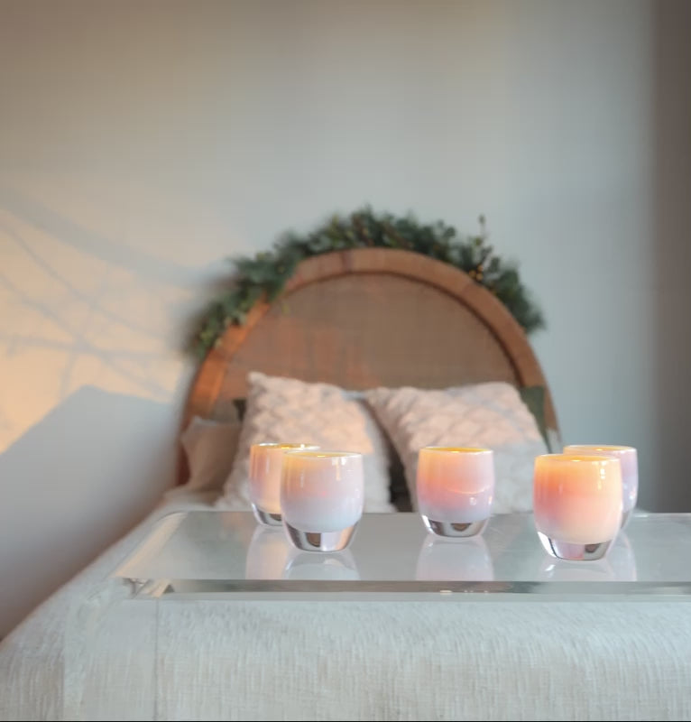 panning video of a grouping of miracle, golden pink hand-blown glass votive candle holders perched on an acrylic table at the end of a bed.
