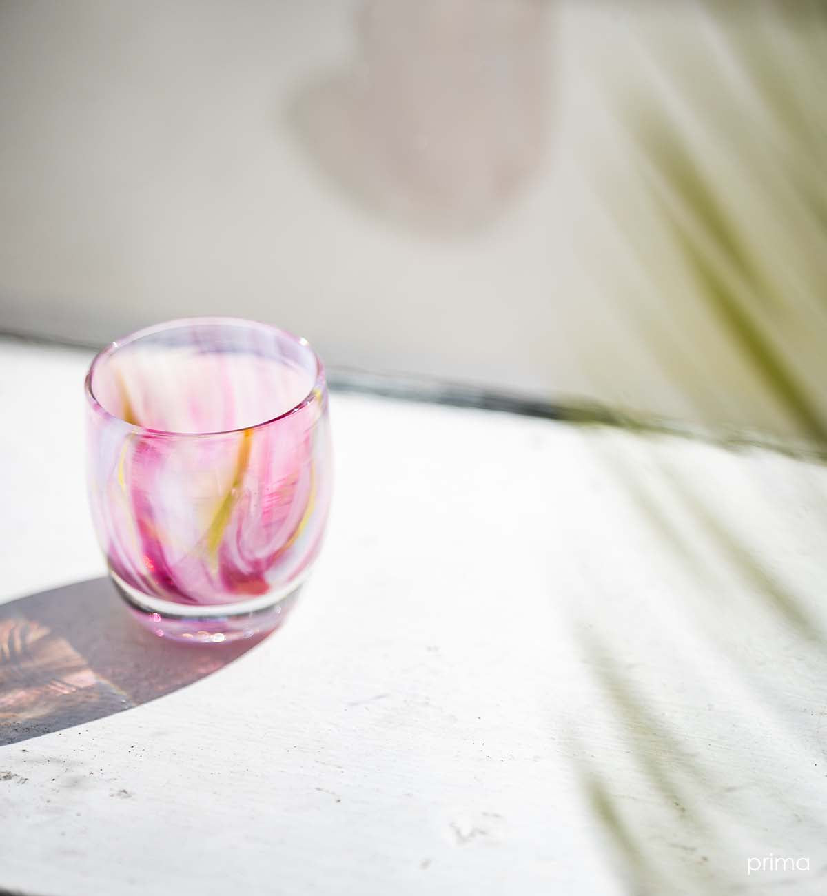 prima pink multi-colored petal, hand-blown glass votive candle holder on a cream surface sitting in the sunshine.