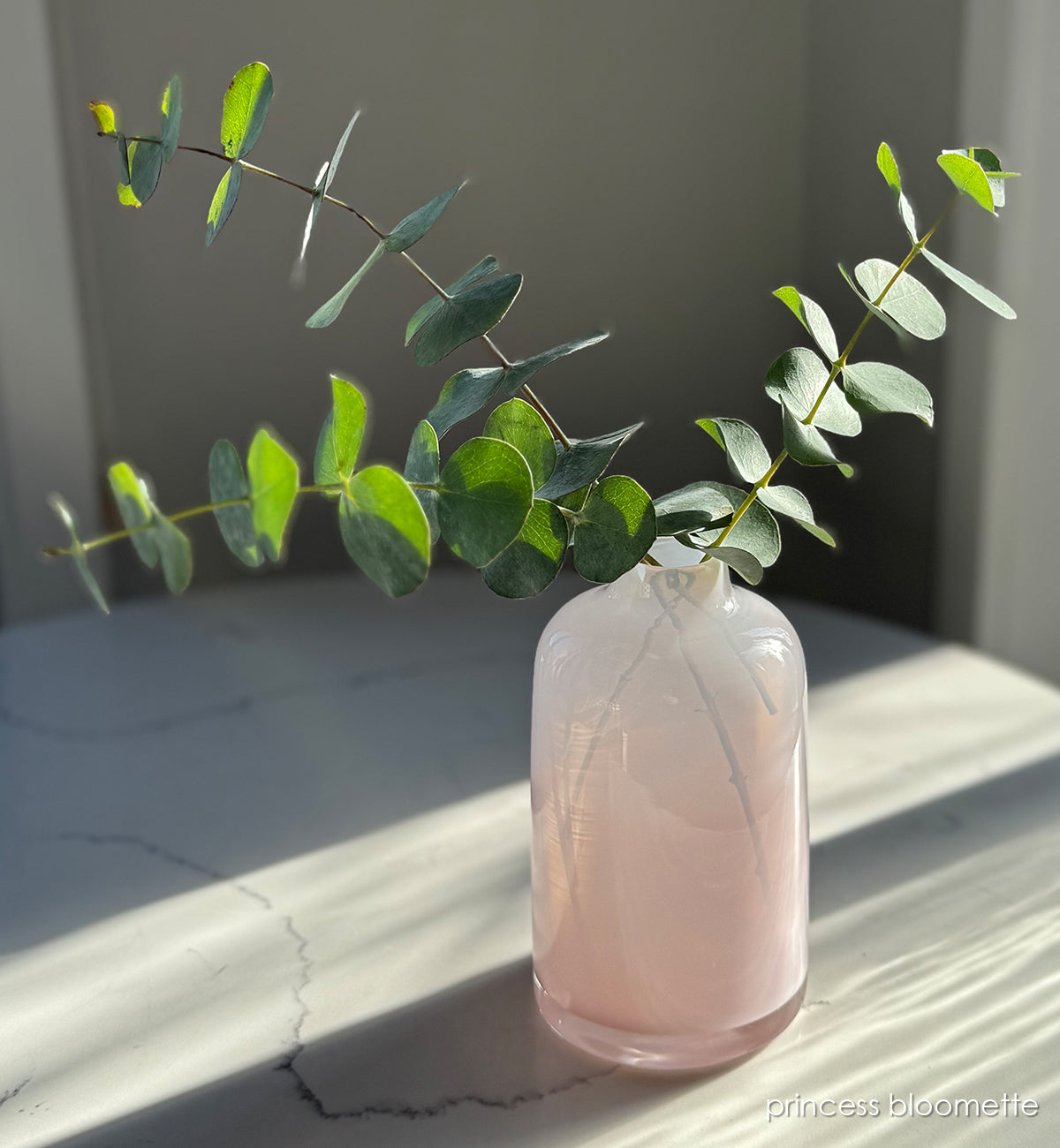 'priness bloomette'  light pink opaque hand-blown glass flower bud vase holding green eucalyptus plant on white and grey marble table. 