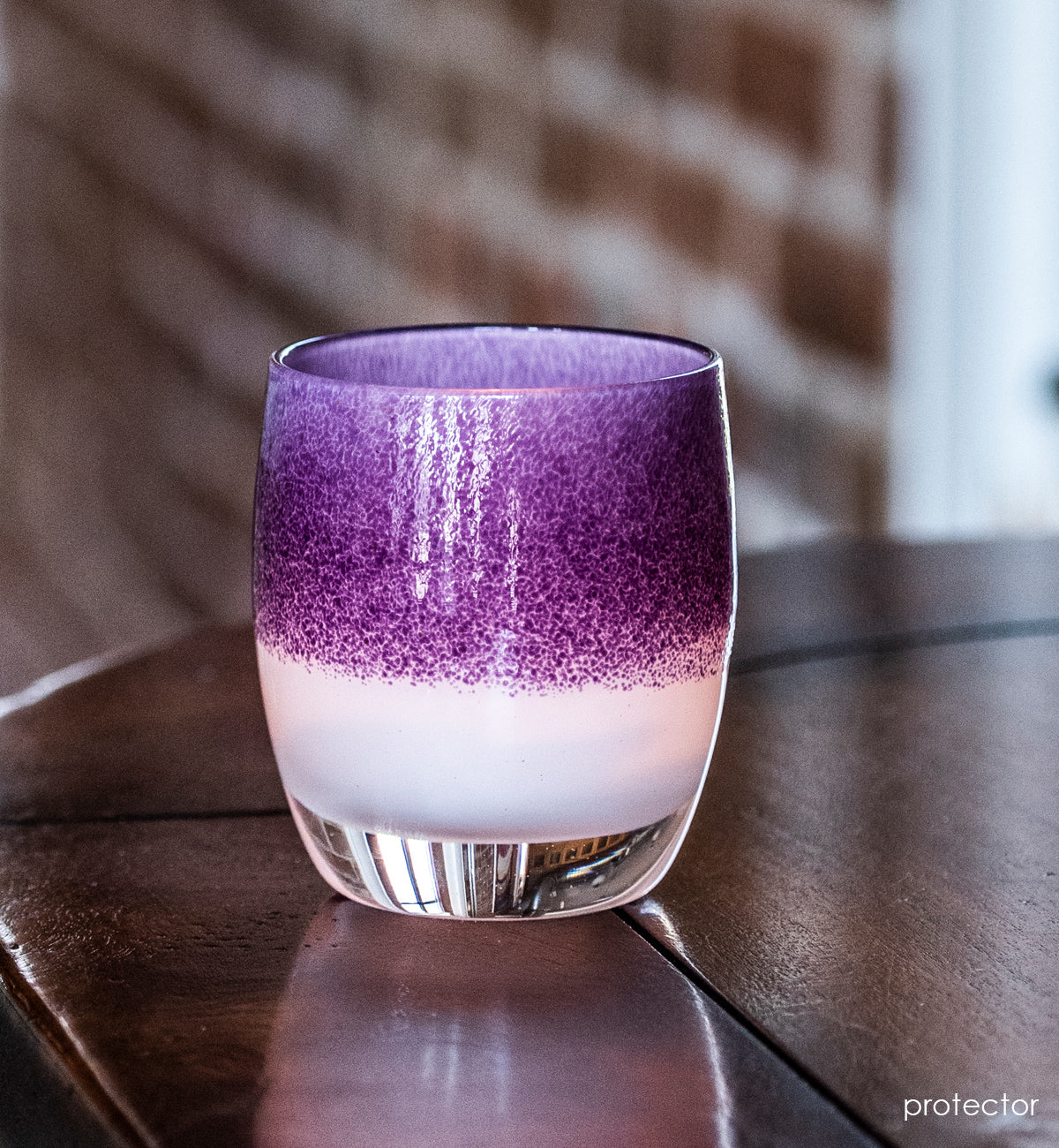 protector purple on white, hand-blown glass votive candle holder, sitting on a dark wood counter backed by exposed brick.