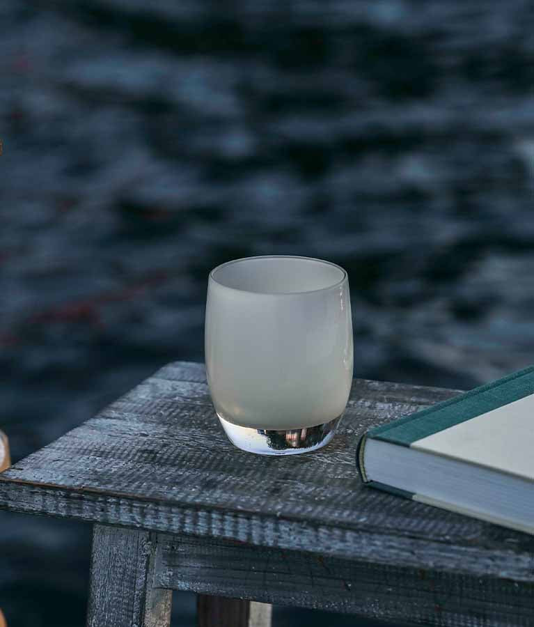 quietude cream hand-blown glass votive candle holder.Sitting near the water with a book and chair.