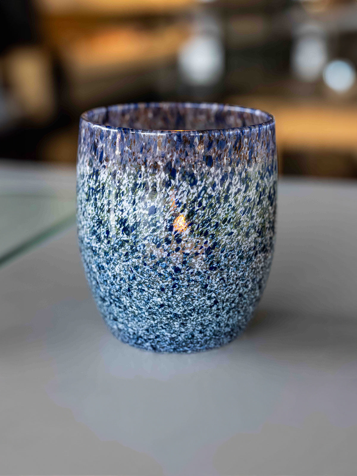 Speckled blue, green and white glass candle holder on a reflective surface with a blurred background
