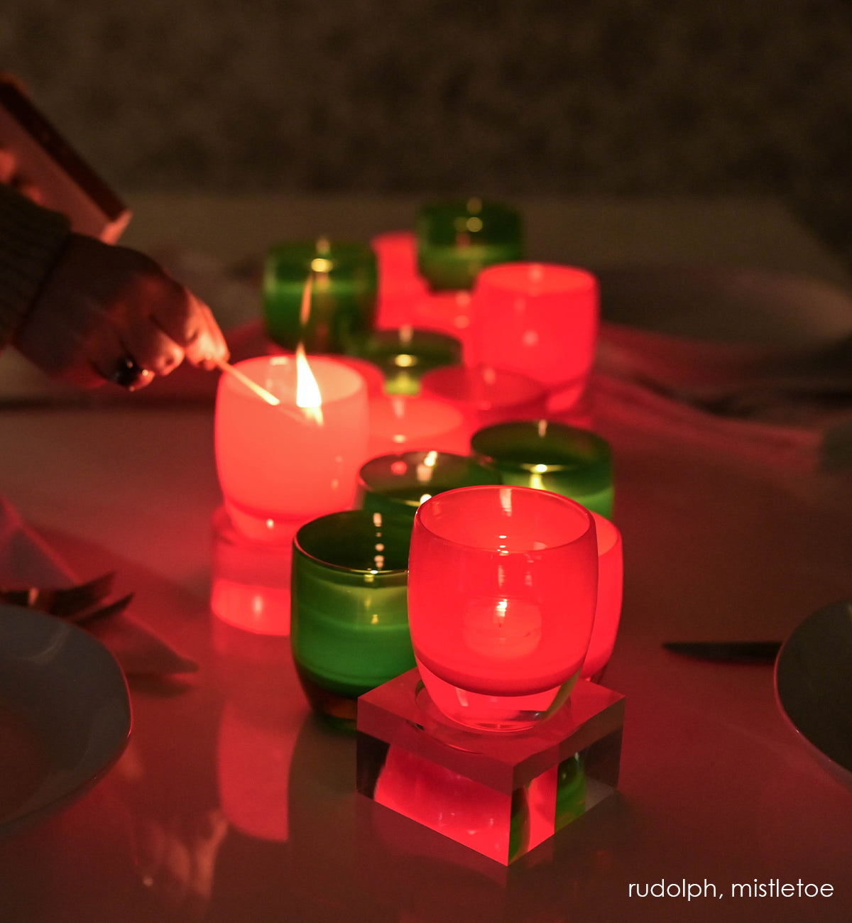 several rudolph velvet red hand-blown glass votive candle holders lit on a table with mistletoe and dinner plates.