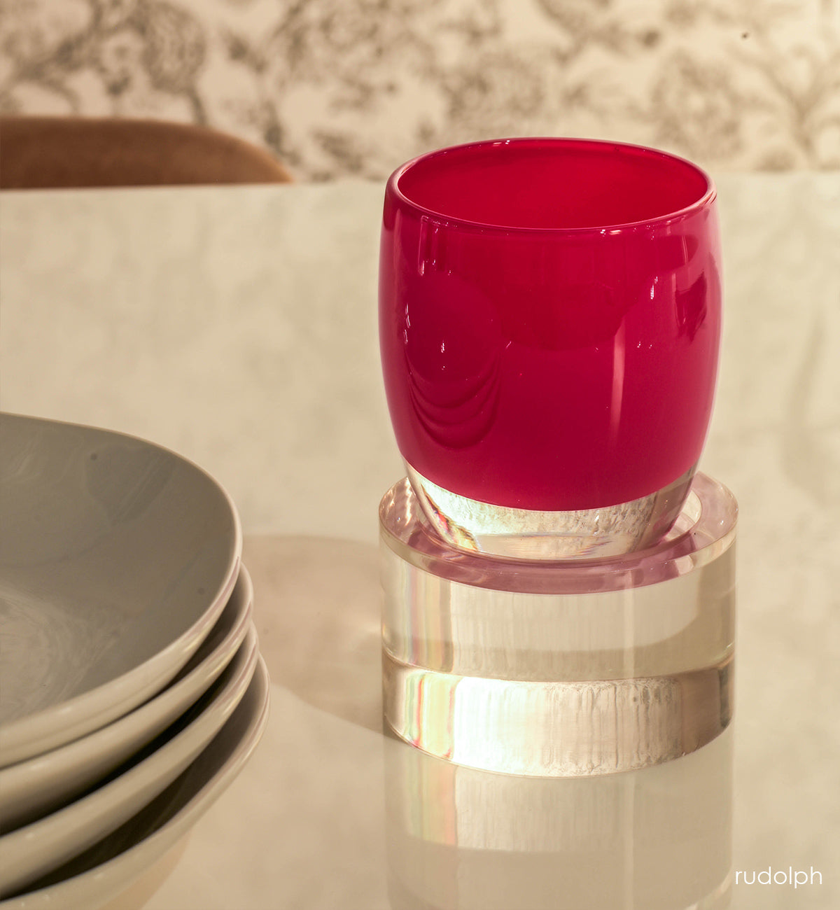 rudolph velvet red hand-blown glass votive candle holder on a square baby stand on a cream dining table next to plates.