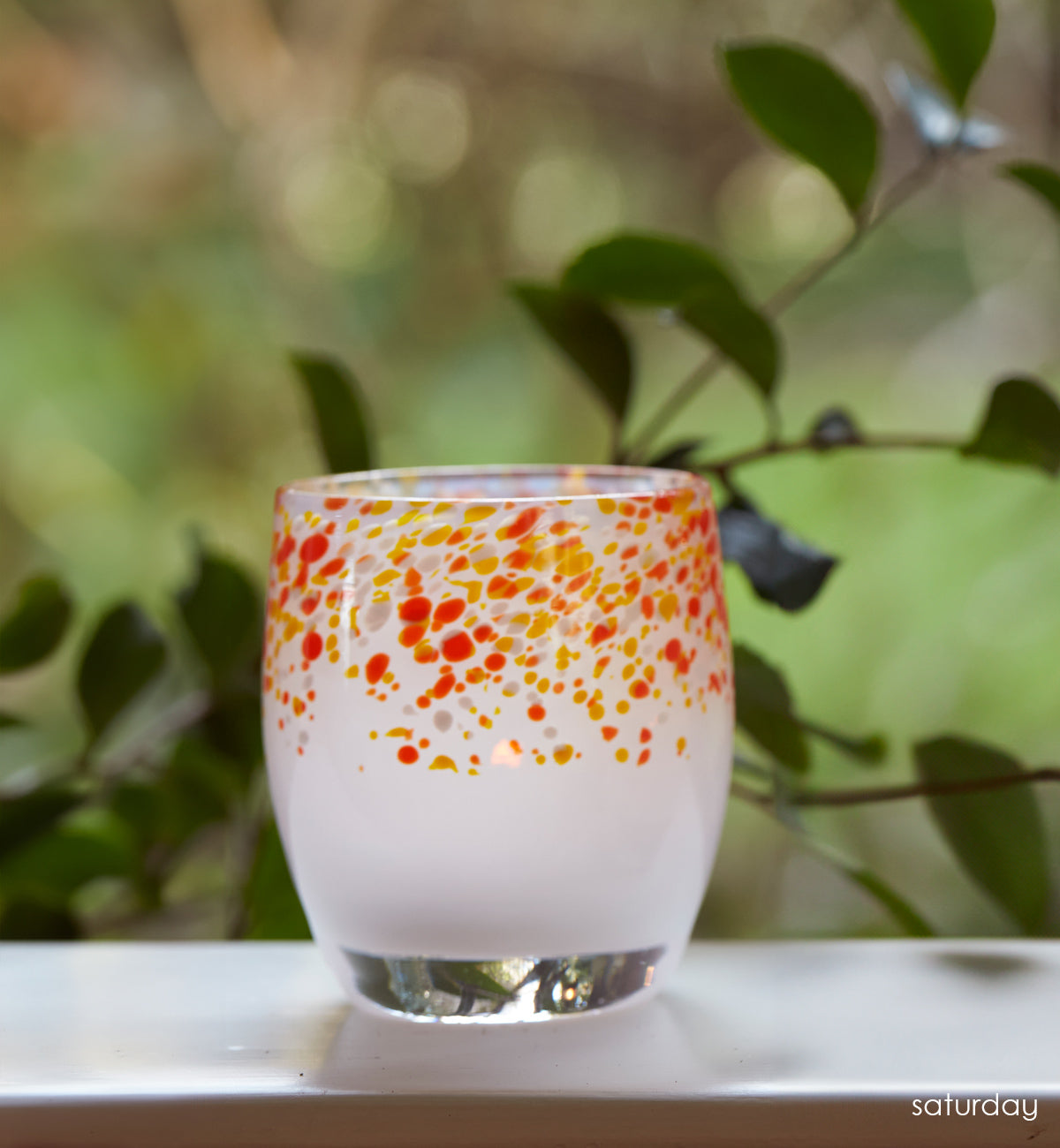 saturday red and orange frit topped white hand-blown glass votive candle holder, on a windowsill looking over a green yard.