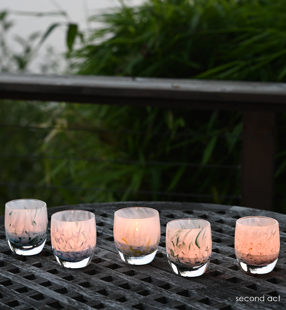 group of five second act hand-blown glass votive candle holders made from recycled glass from 43 colors on a wood patio table with greenery in background.