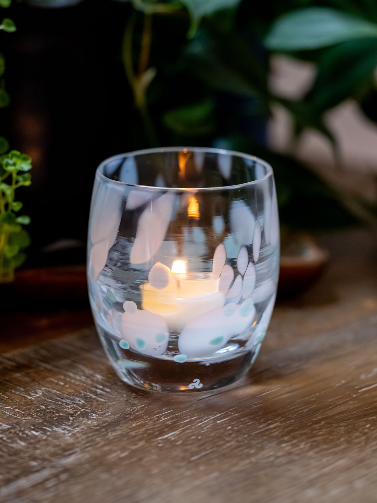 Clear glass candle holder with white and blue spotted design containing a lit candle on a wooden surface, blurred greenery in the background.