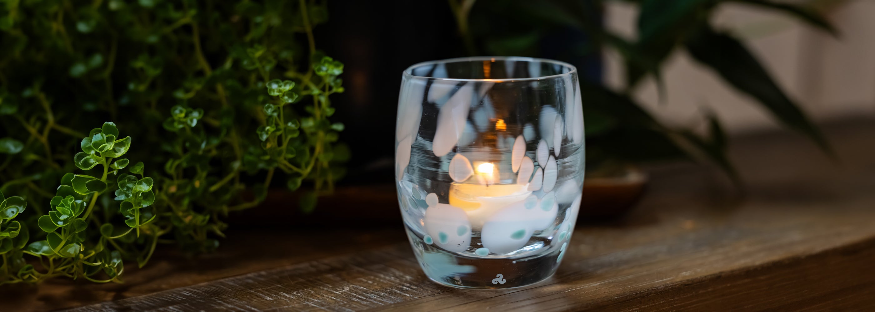 Clear glass candle holder with white and blue spotted design containing a lit candle on a wooden surface, blurred greenery in the background.