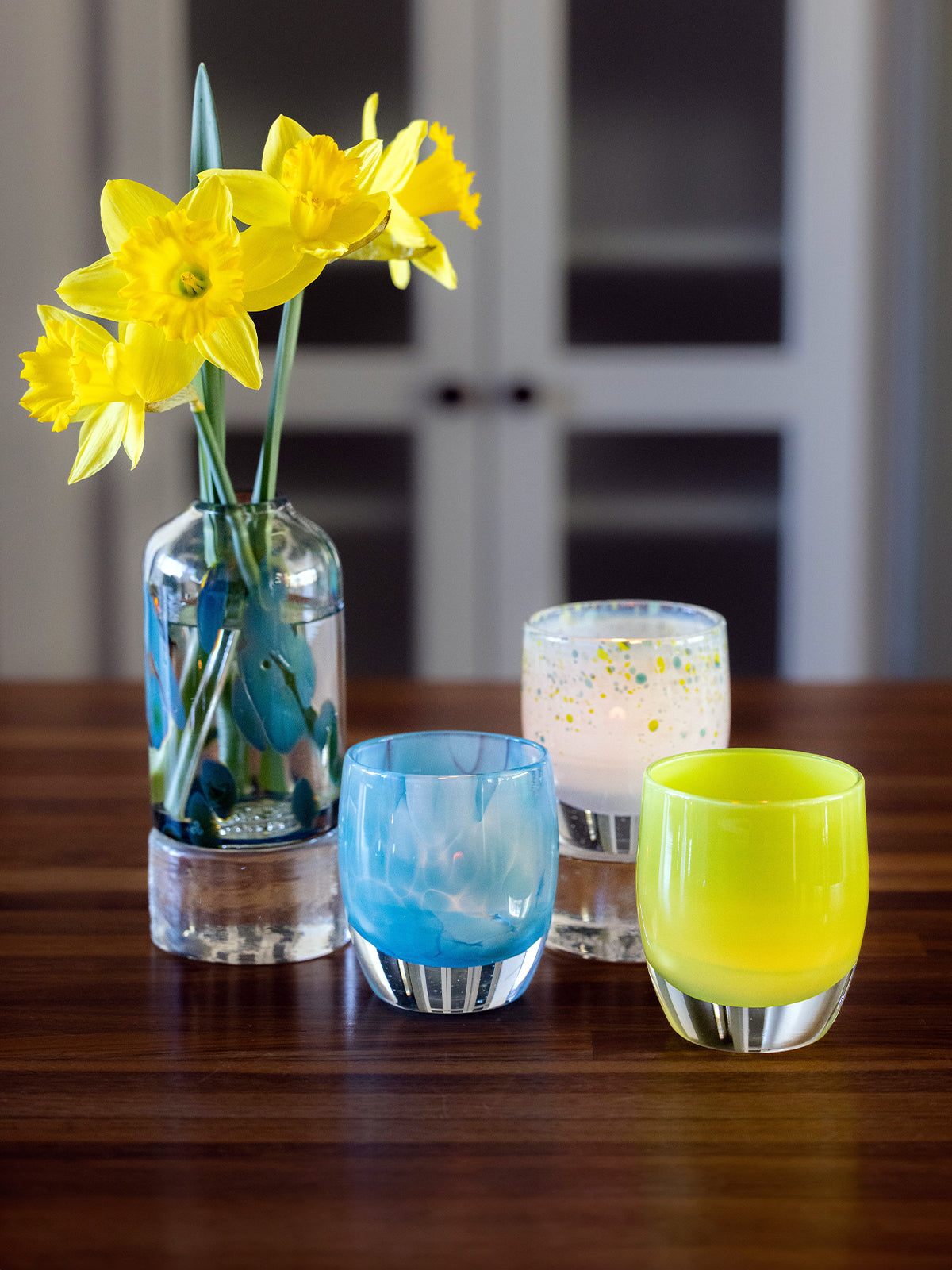 colorful spring glass candle holders and bloomette vases with yellow flowers on a wooden table.