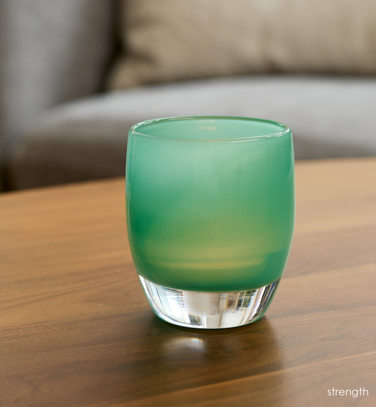 strength teal green hand-blown glass votive candle holder on a wood coffee table with gray couch in background.