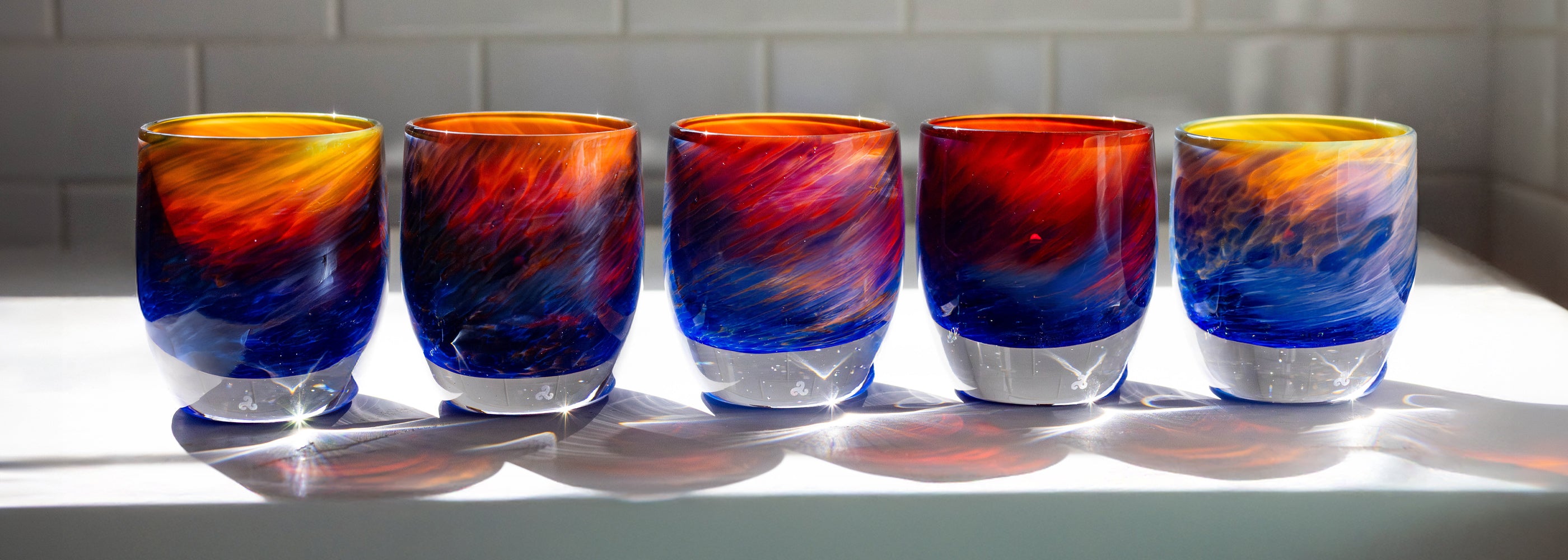 group of 'summer sunset' glassybaby red, blue, yellow hand-blown glass candle holder sitting on white counter top with white tile in the background.