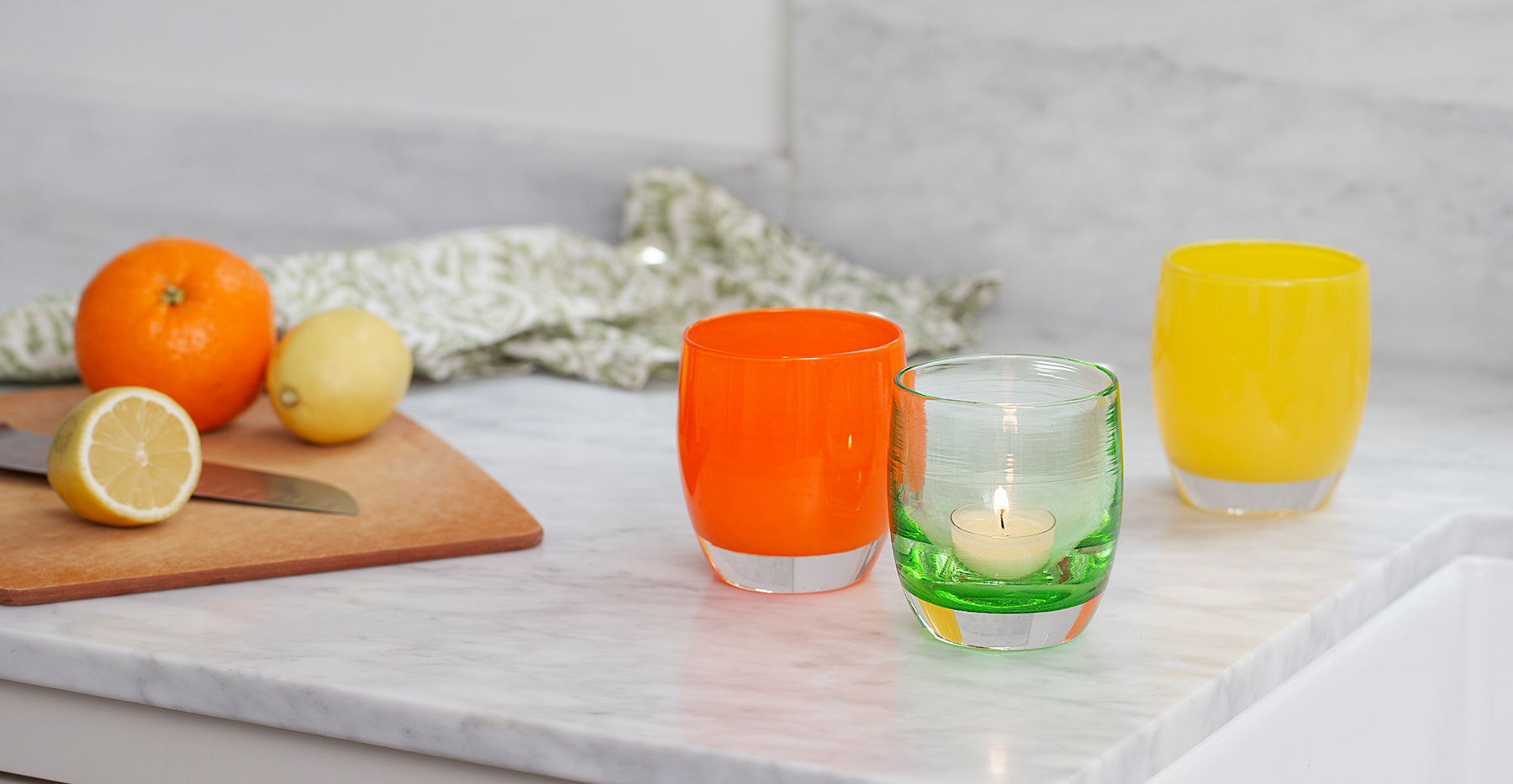 tangerine, bright orange hand crafted glass candle holder paired with clover, transparent green hand-blown glass candle holder and sunshine, an opaque yellow hand-blown glass candle holderon a marble table top.