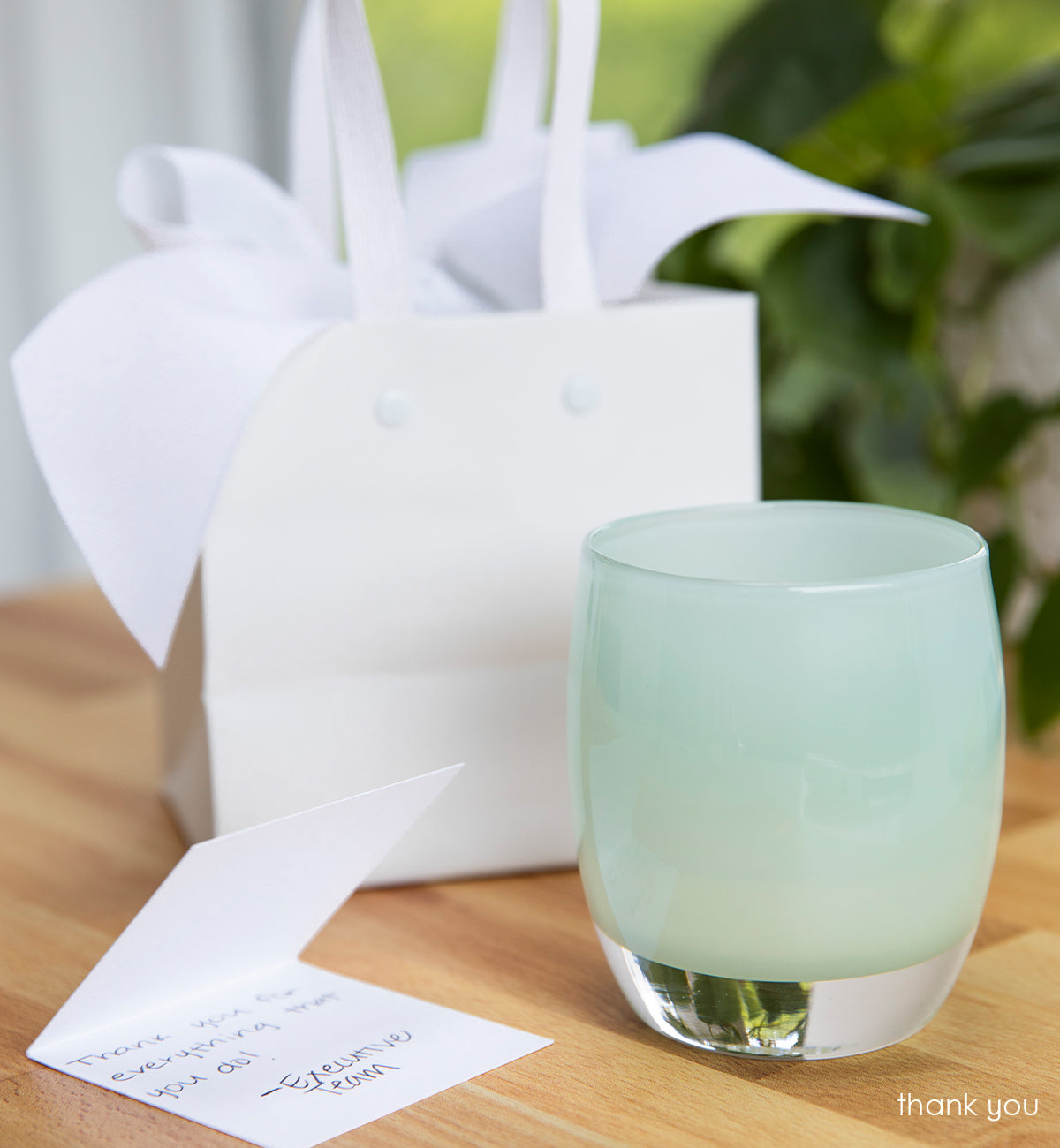 thank you mint green hand-blown glass votive candle holder on light wood table next to a notecard and glassybaby packaging. blurred green plant in background.