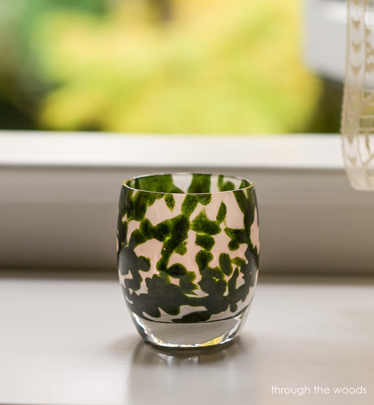 through the woods hand-blown dark green with glitter glass votive candle holder on a surface in front of an open window with lace curtains.
