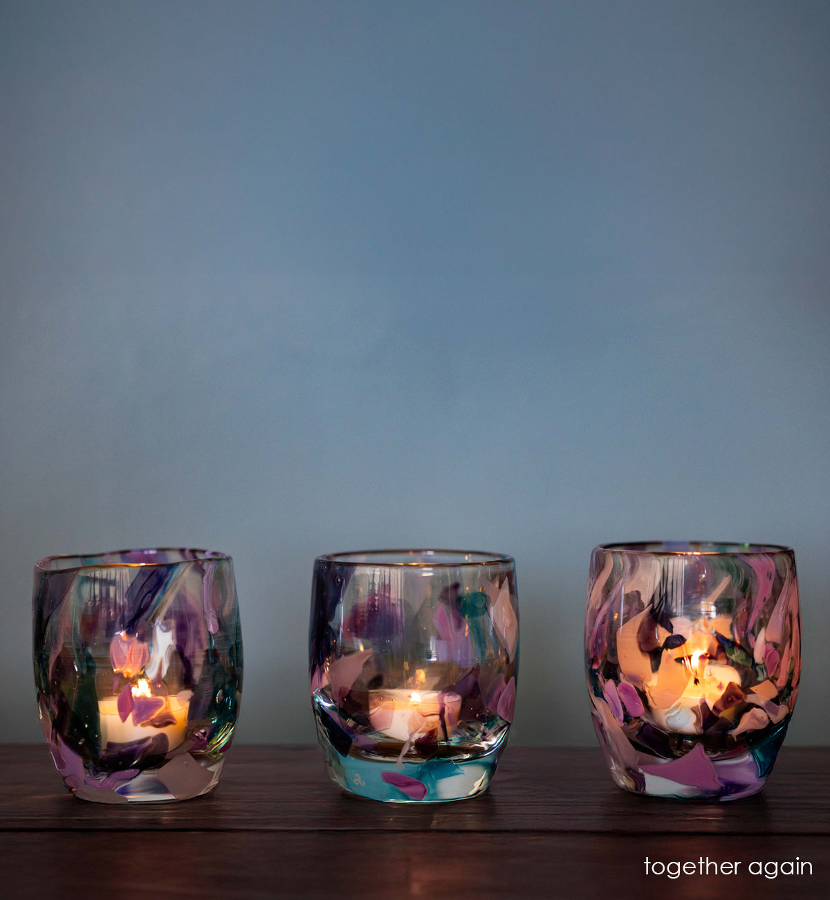 three lit 'together again' multi-color (blue, purple, brown, light blue) hand-blown glass candle holders on wood table.