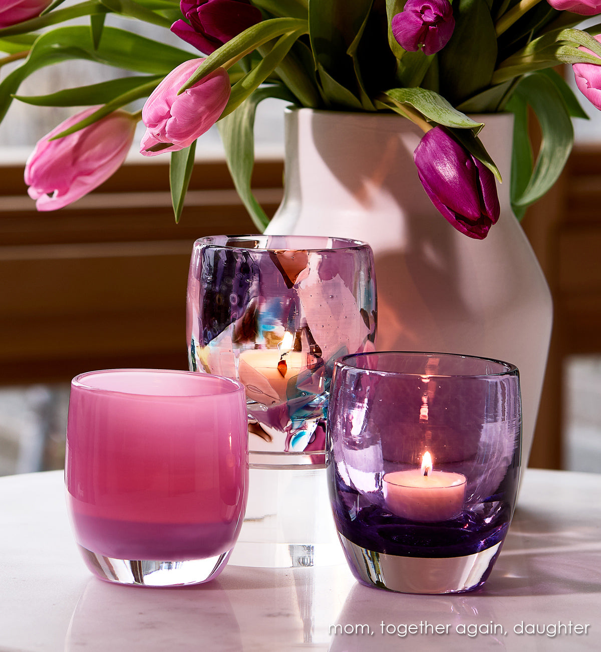 'mom', 'together again', 'daughter' multi-color (blue, purple, brown, light blue) hand-blown glass candle holder on white table with vase of pink and purple tulips in the background.