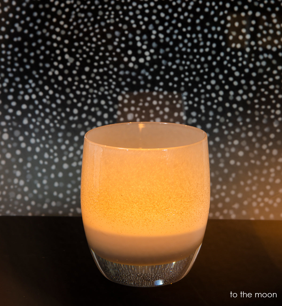 to the moon light grey and white, hand-blown glass votive candle holder on dark surface with a starry night background.