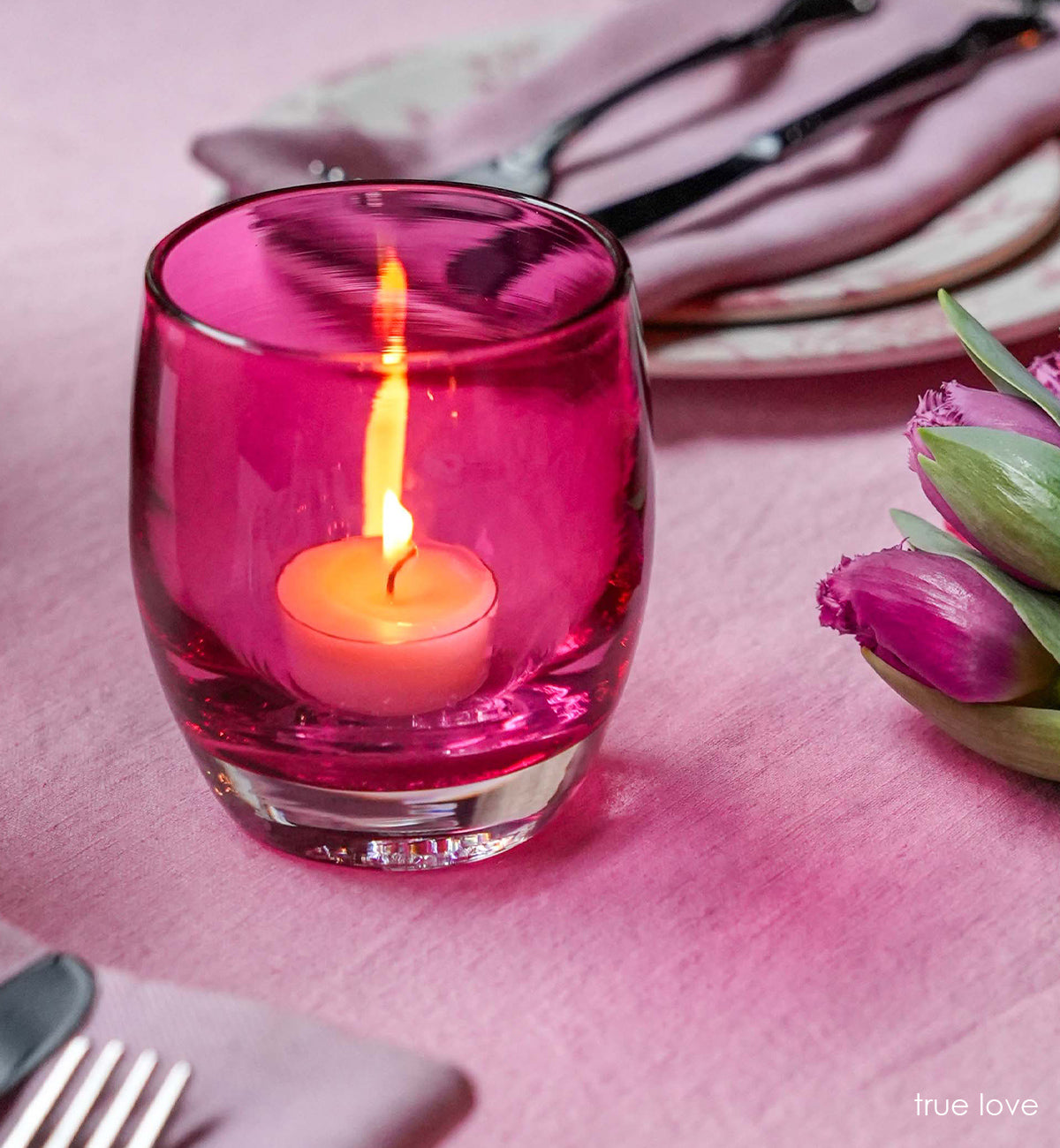true love, transparent deep raspberry pink, hand-blown glass votive candle holder on a pink table cloth next to flowers and place settings.