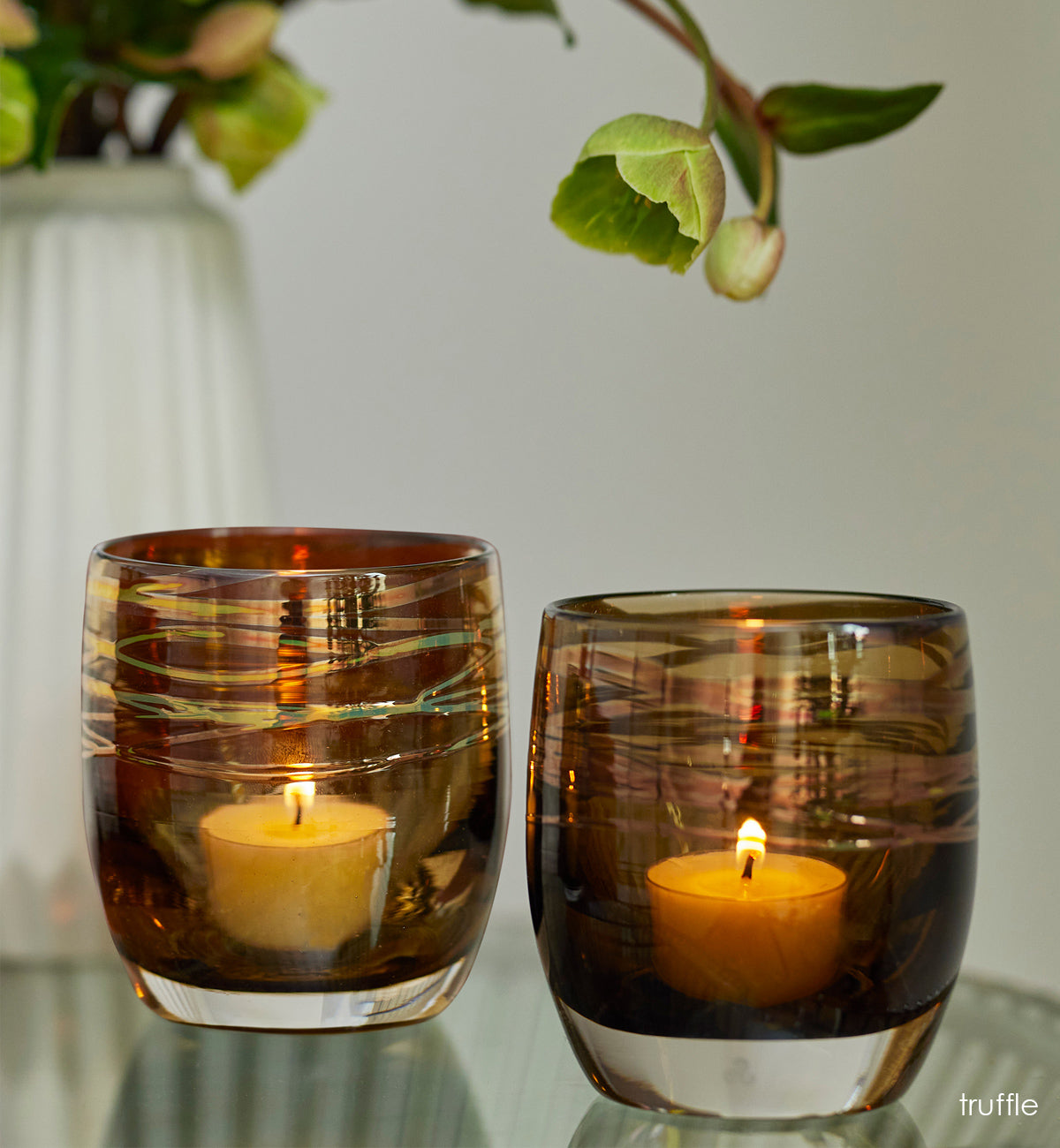 grouping of truffle, brown with metallic shimmer wrap hand-blown glass votive candle holders lit on a glass table. green plant in white vase in background.