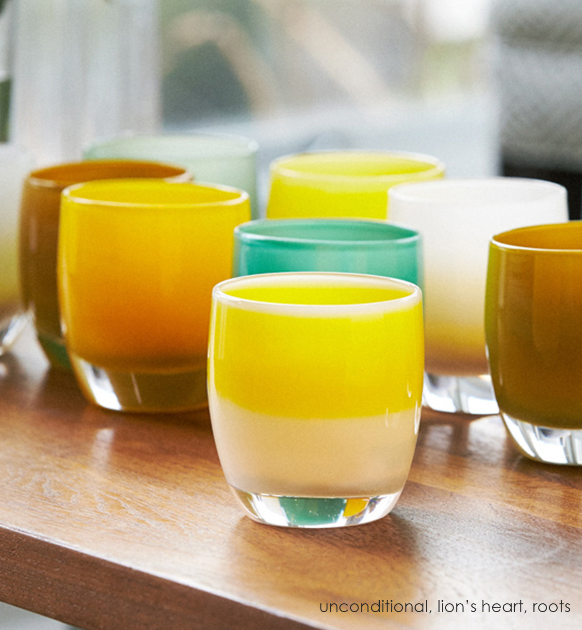 unconditional, white with yellow stripe, hand-blown glass votive candle holder on a light wood table with grouping of lion's heart and roots and peaceful.