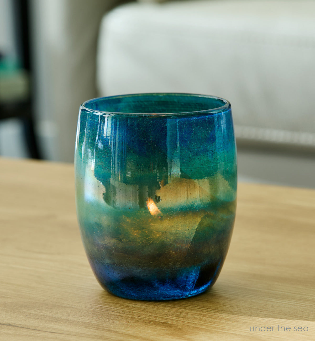under the sea, blue green luster hand-blown glass votive candle holder with a tealight lit inside, and the flame peaking through the translucency. Placed on a light wood coffee table.
