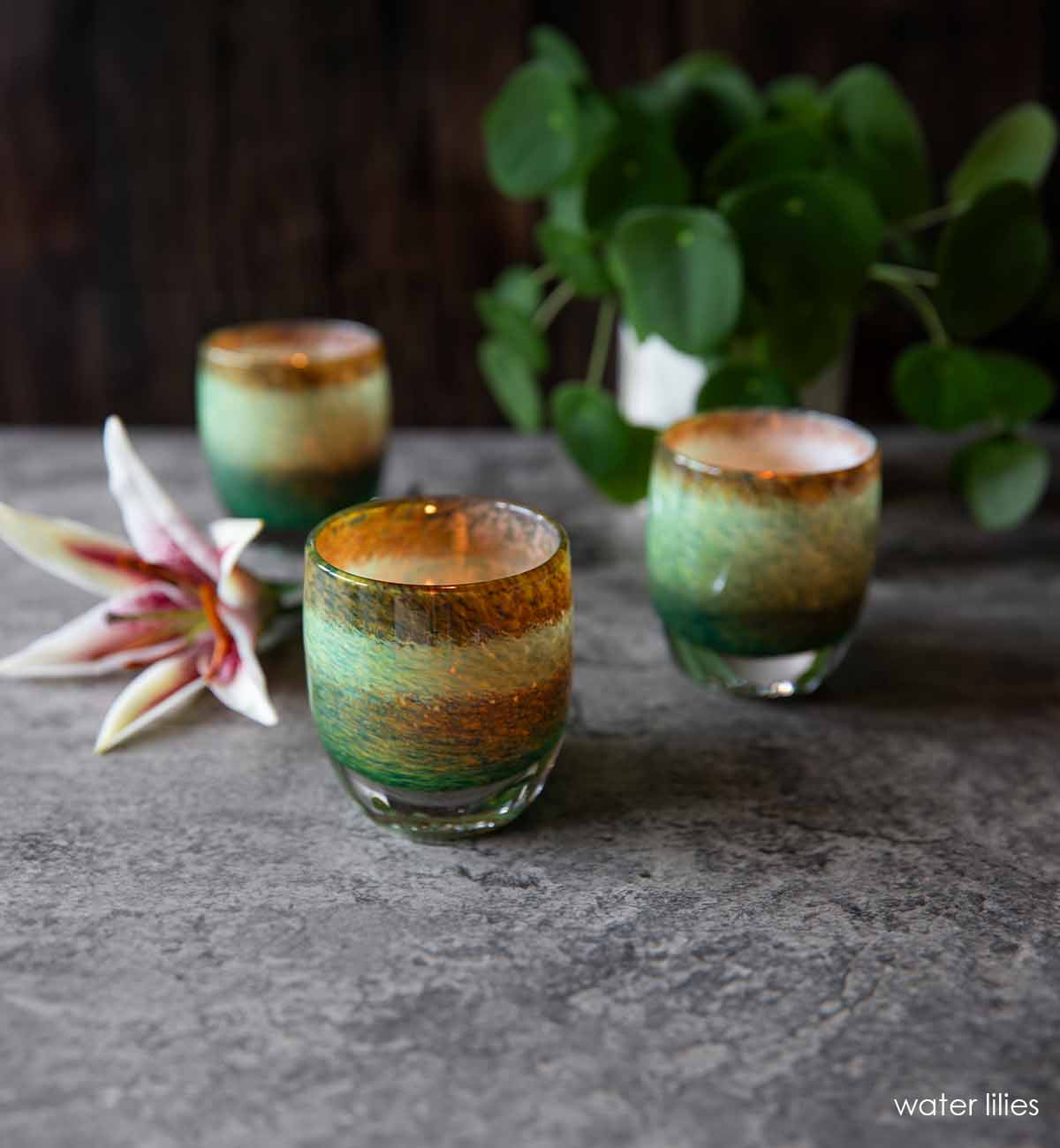 Three pond colored water lilies votive candle holders lit on a stone table with a pilea plant and a lily flower.