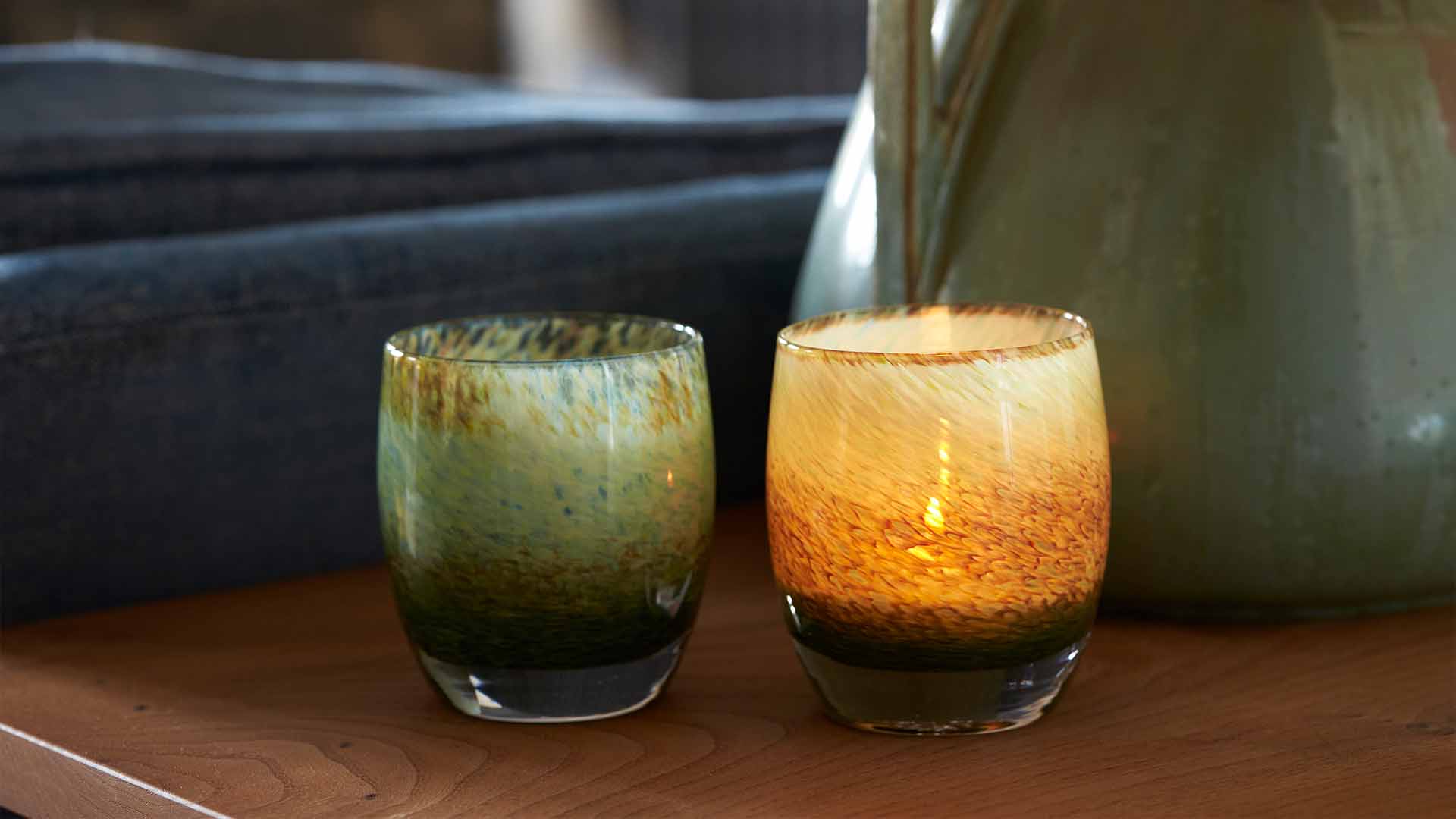 a pair of water lilies sit on a wooden side table, one glowing with a warm light. garden pond earthy green blue textured hand-blown glass votive candle holder.