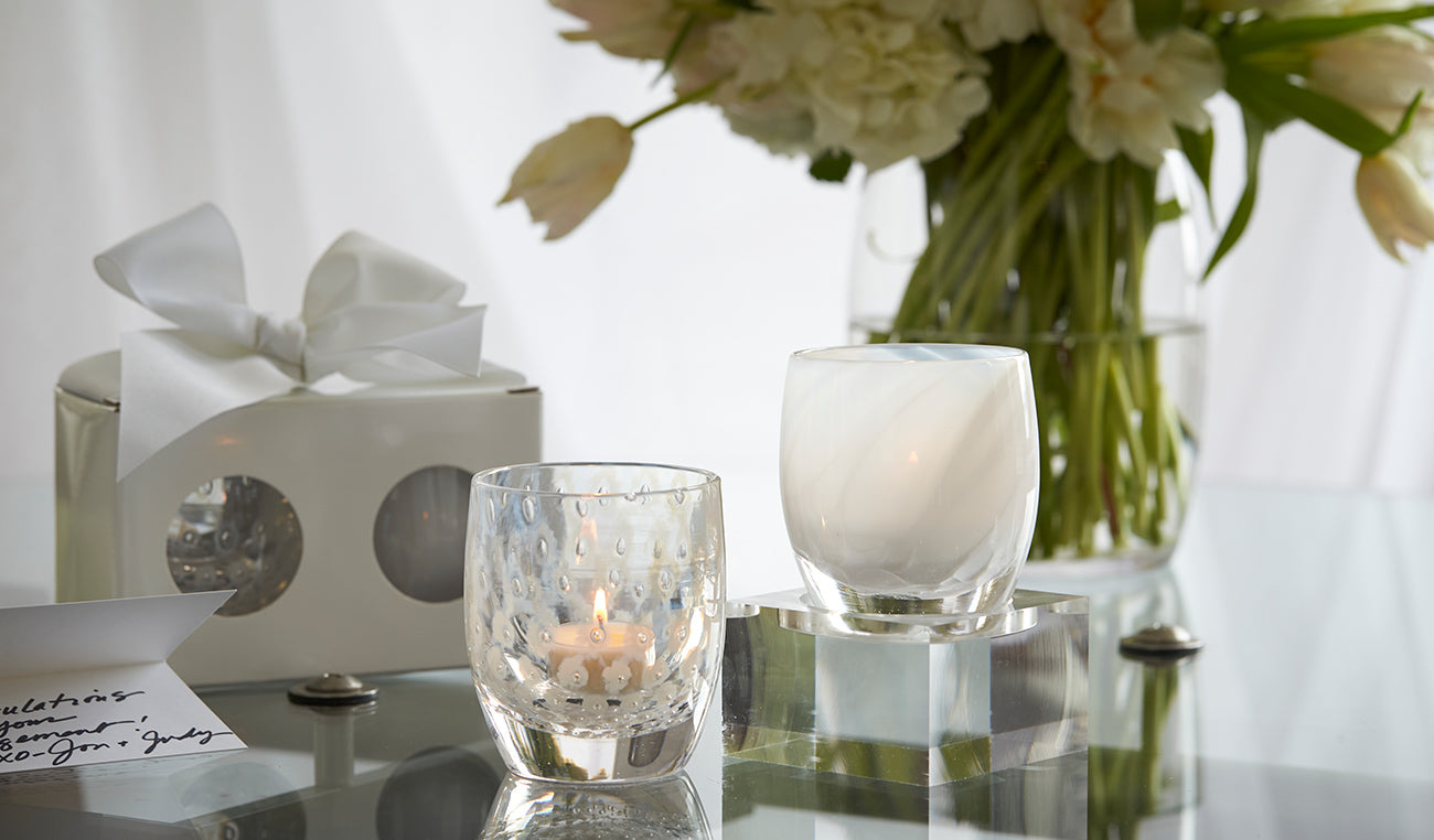 become one set includes a yes glassybaby and an embrace glassybaby - both white patterned hand-blown glass candle holders sitting on a glass table with flowers in the background.