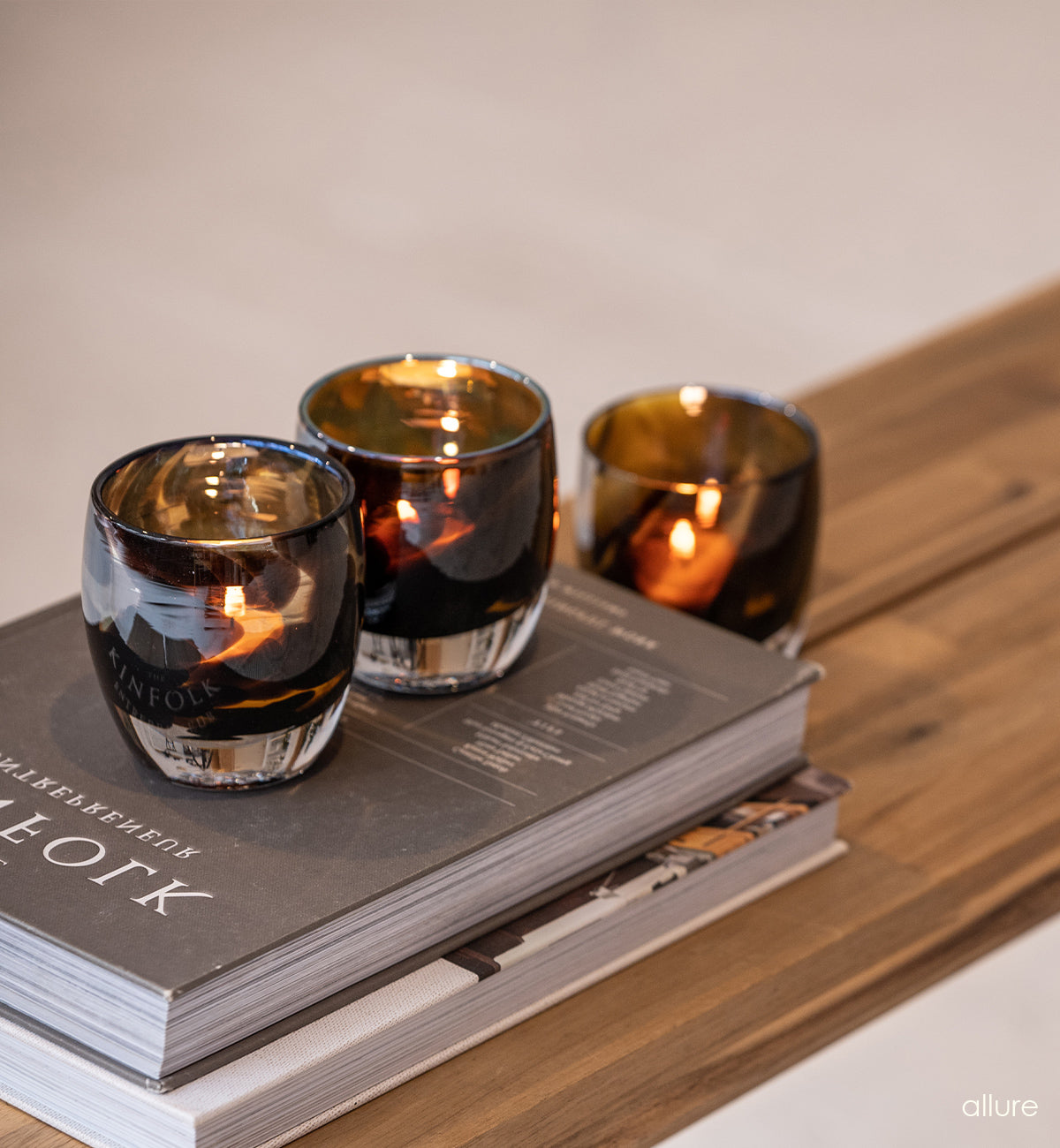 three allure, brown and gold, hand-blown glass votive candle holders on coffee table books and wood surface.