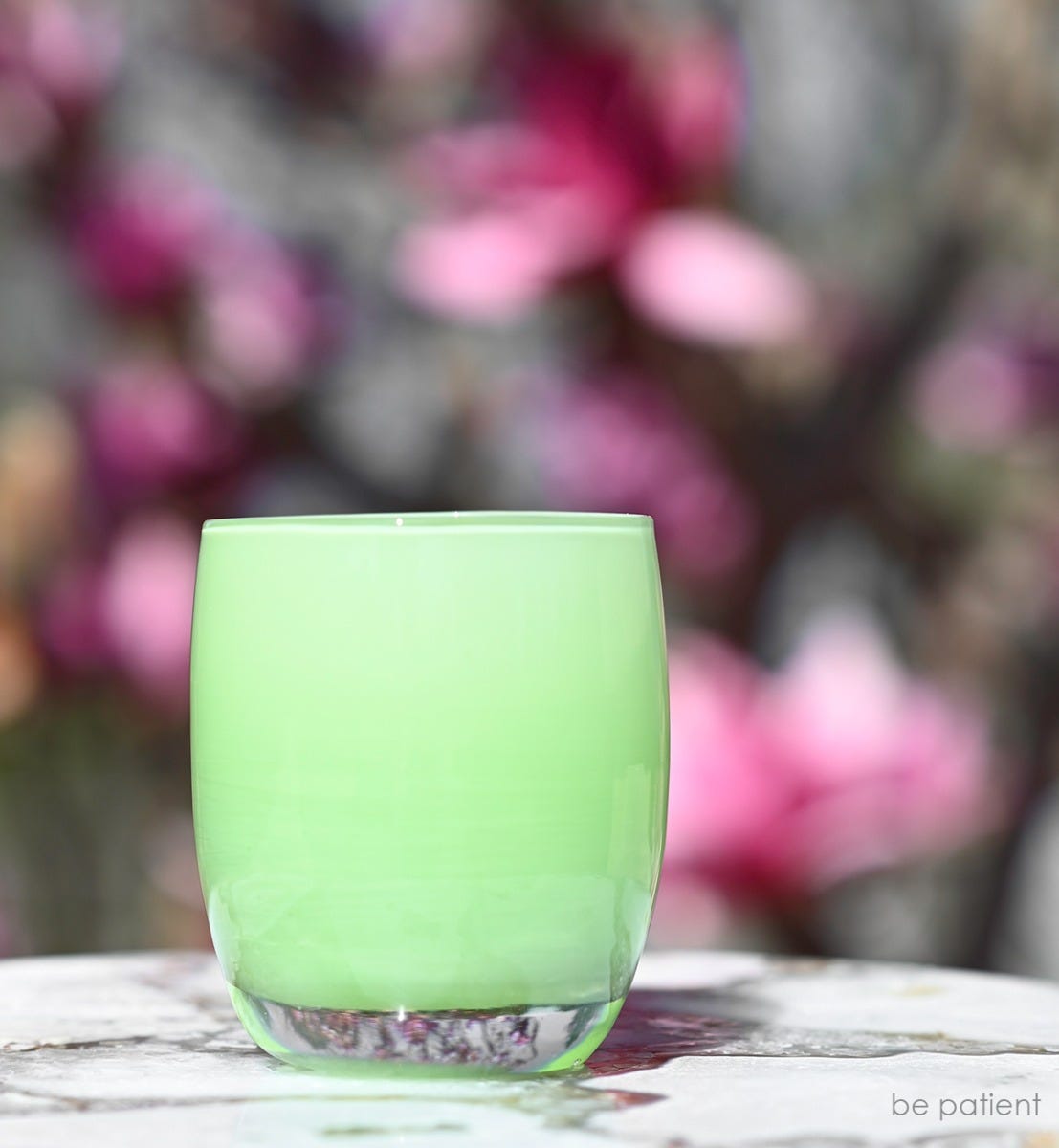 be patient, light green hand-blown glass votive candle holder on marbled outdoor table with pink flowers blurred in background.