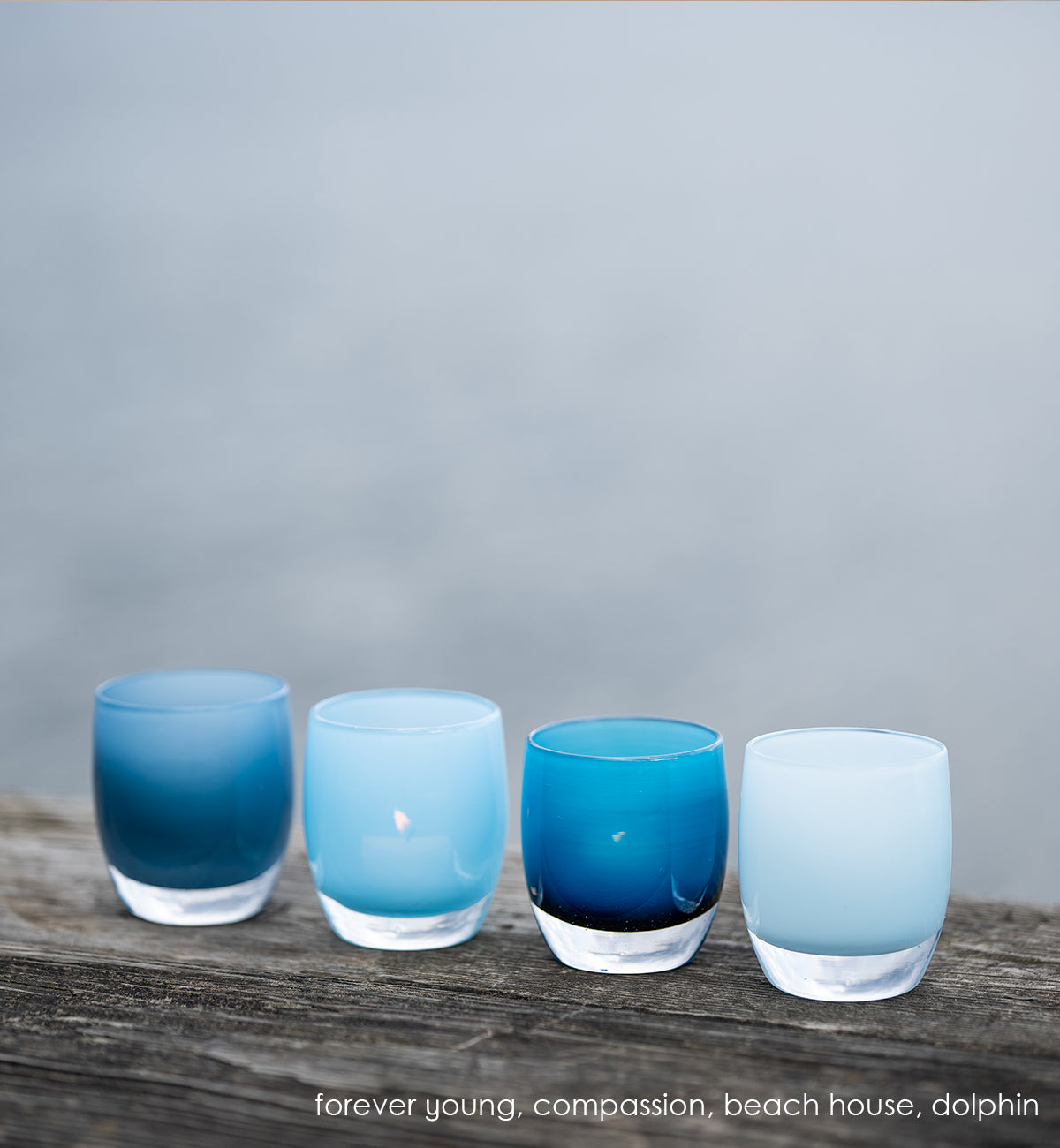compassion powder blue, hand-blown glass votive candle holder in a line up with forever young, beach house and dolphin outside on wood surface.