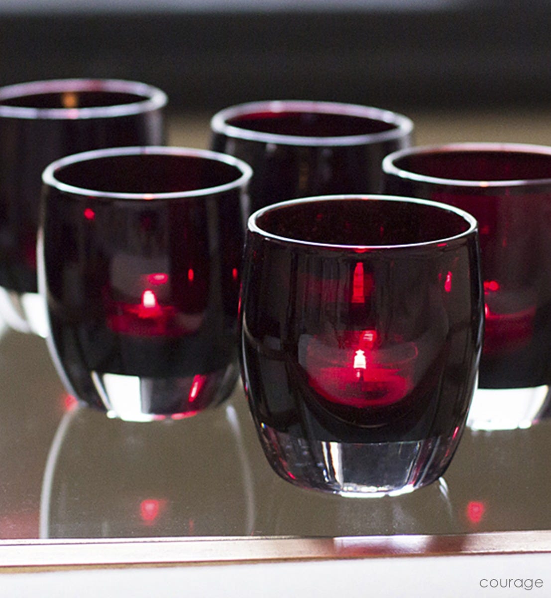 several courage, translucent mahogany red, hand-blown glass votive candle holders lit with tealights on a wood and glass coffee table.